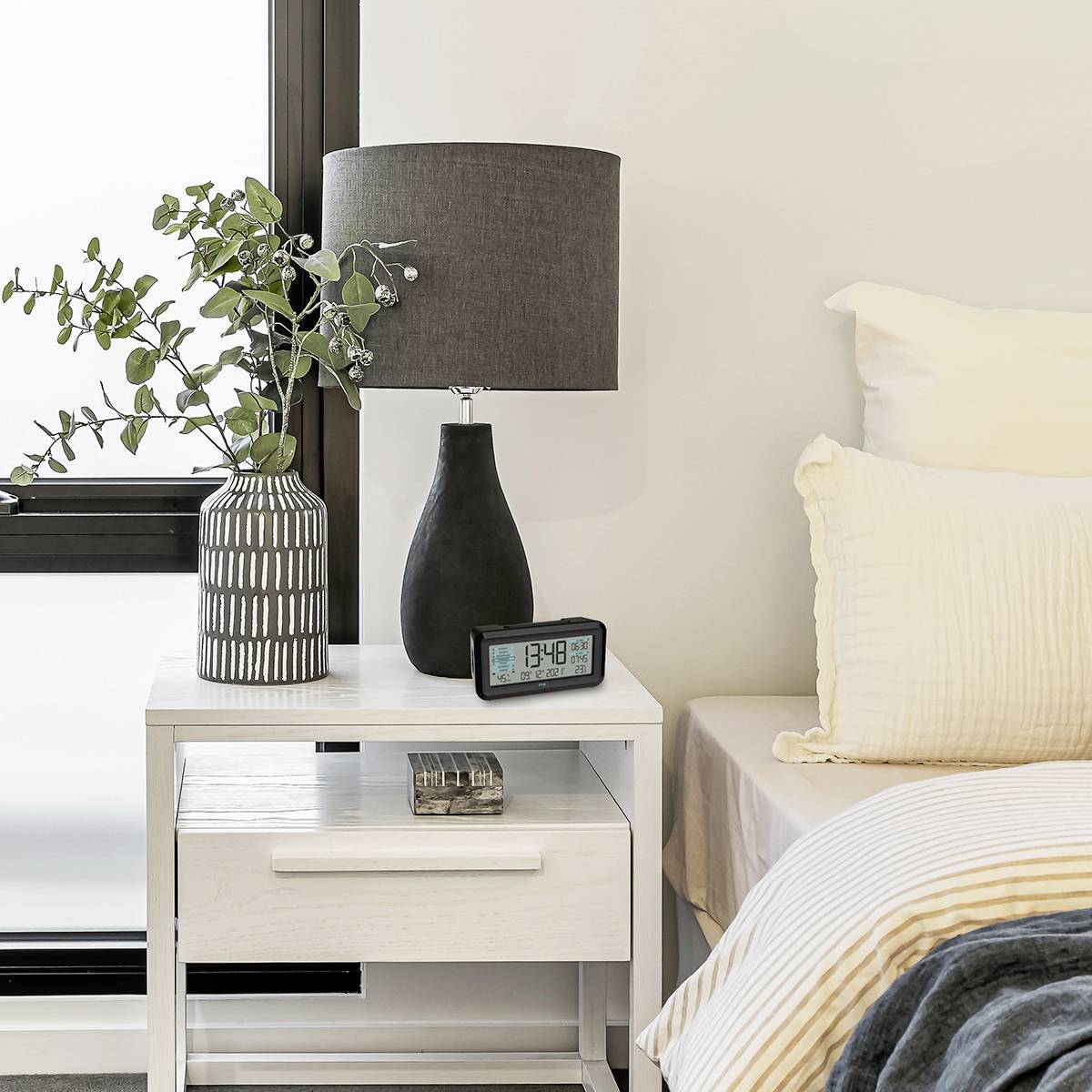 Stolik nocny z cyfrowym zegarem, wskazującym '13:48'. Obok lampka stołowa, wazon z zielonymi gałązkami. Częściowo widoczne fragment łóżka.