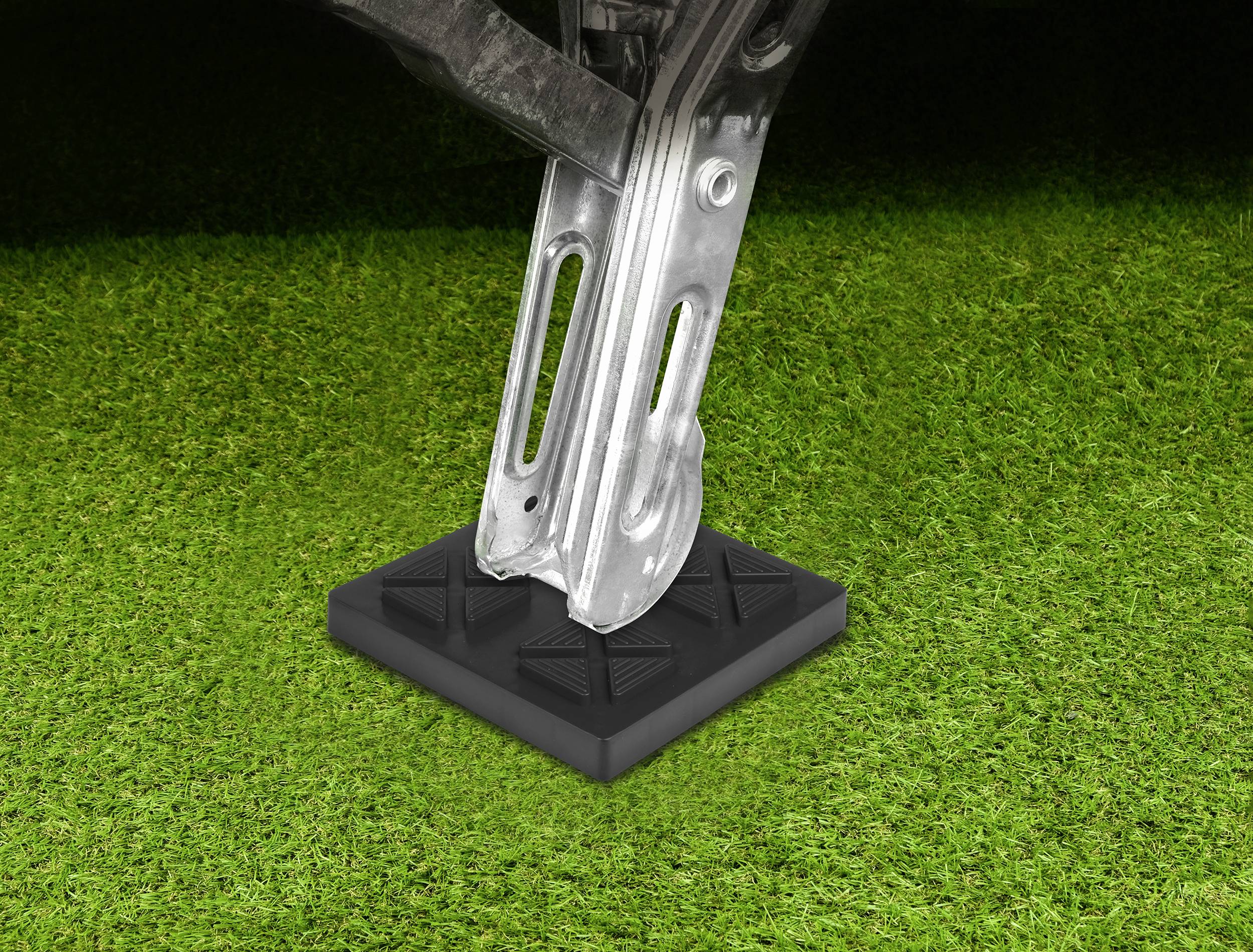 Podnośnik samochodowy spoczywa na czarnym klinie podkładowym na zielonej trawiastej powierzchni. Klin podkładowy stabilizuje podnośnik.