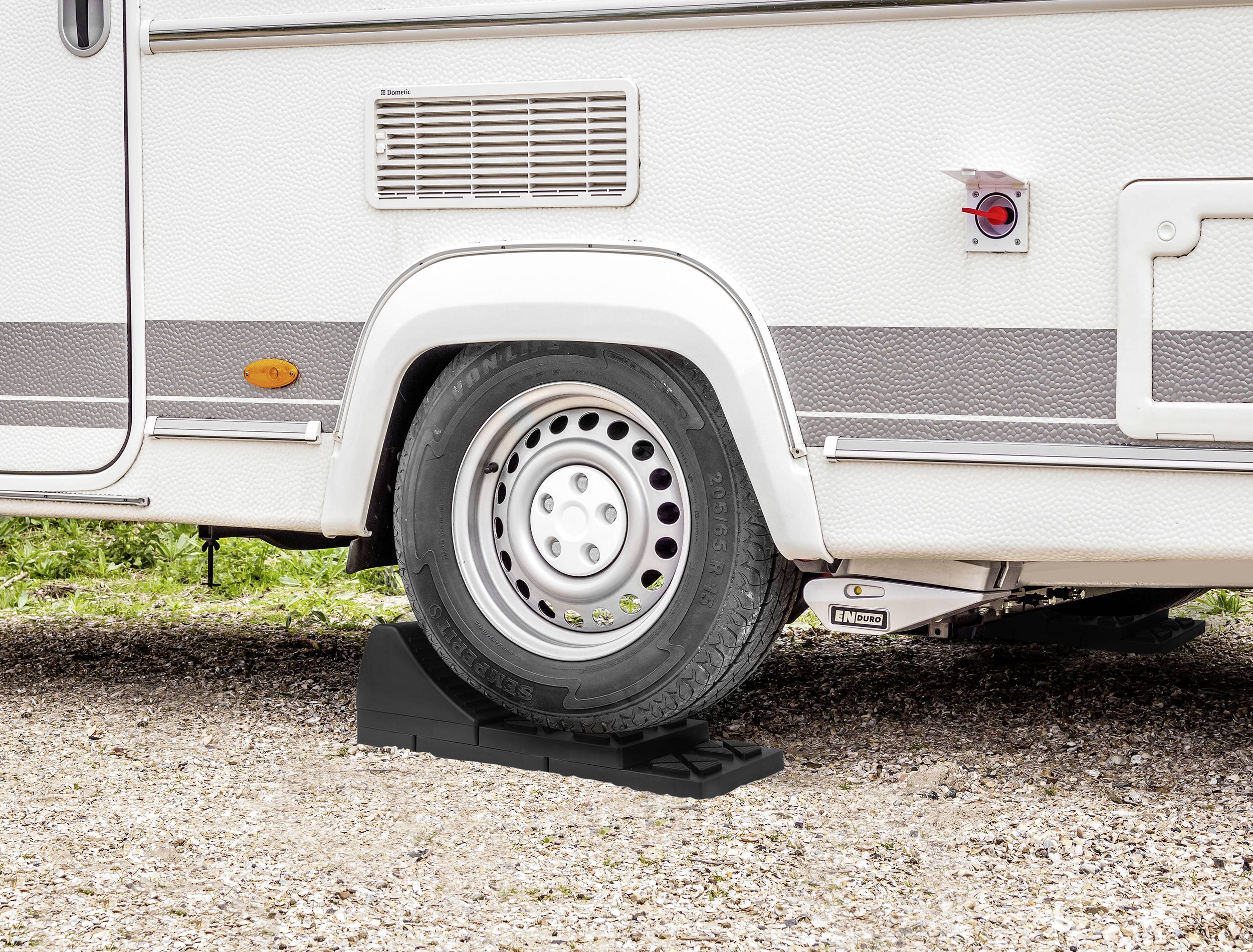 Koło przyczepy kempingowej na czarnym klinie przeciwpoślizgowym, zaparkowane na żwirze. Klin stabilizuje koło, zapobiegając jego toczeniu się.