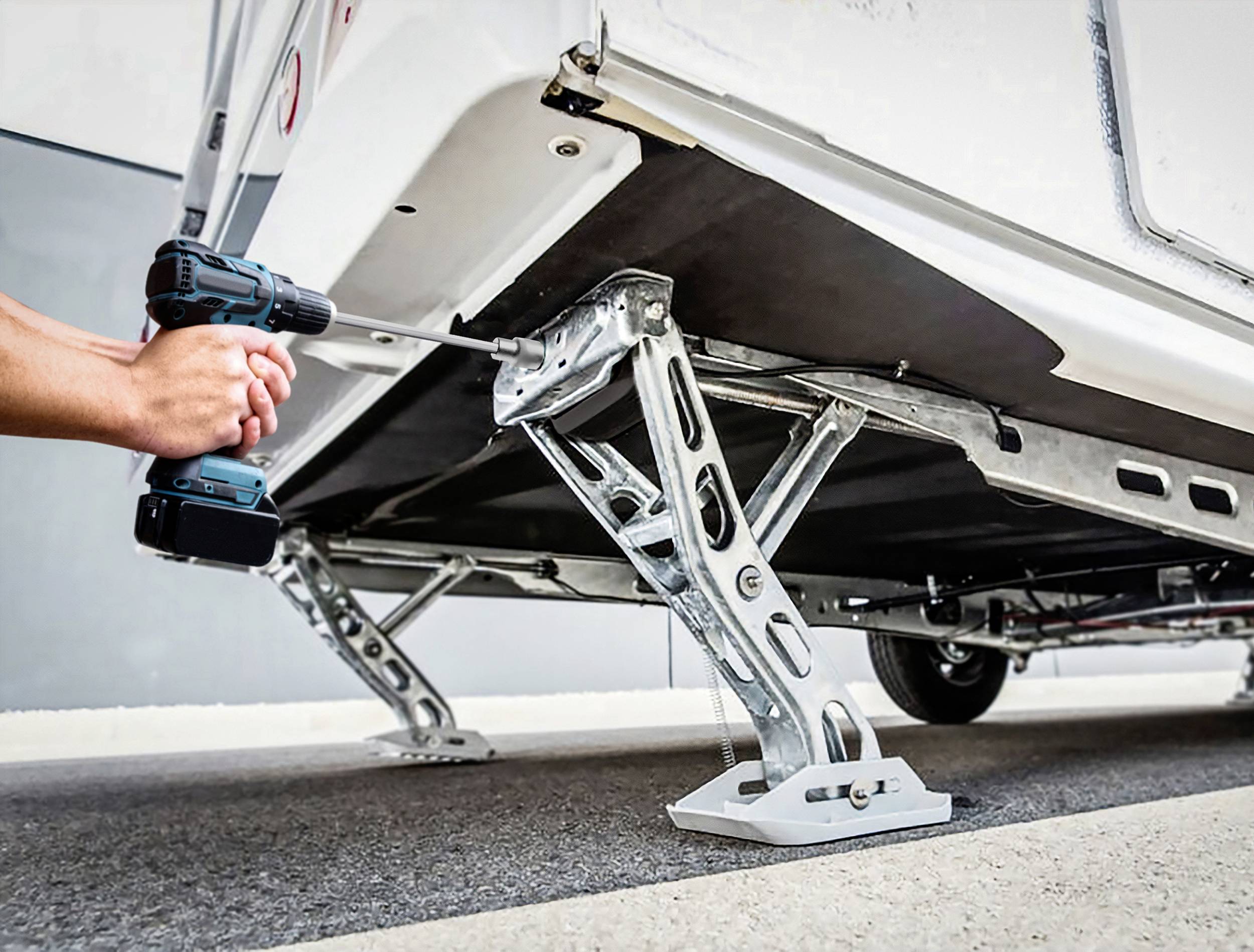 Osoba używa akumulatorowego wkrętarko-wiertarki do wyrównania podpór przyczepy kempingowej. Scena przedstawia precyzyjną pracę ręczną.