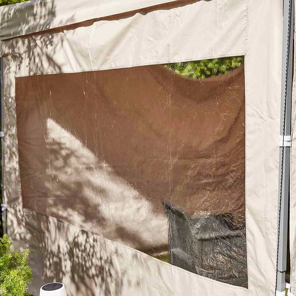 Beżowy pawilon z przezroczystą częścią okienną, jasno oświetlony promieniami słońca. Cienie liści są widoczne na tkaninie.