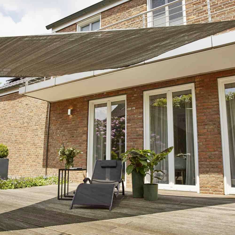 Nowoczesny taras z drewnianym pokładem, leżakiem, doniczkami z roślinami oraz prostokątnym żaglem przeciwsłonecznym. W tle dom z dużymi oknami.