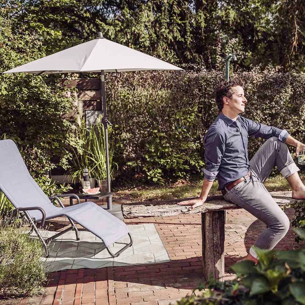 Mężczyzna siedzi zrelaksowany na drewnianym stole w ogrodzie z leżakami i parasolem. Jest słoneczny dzień.