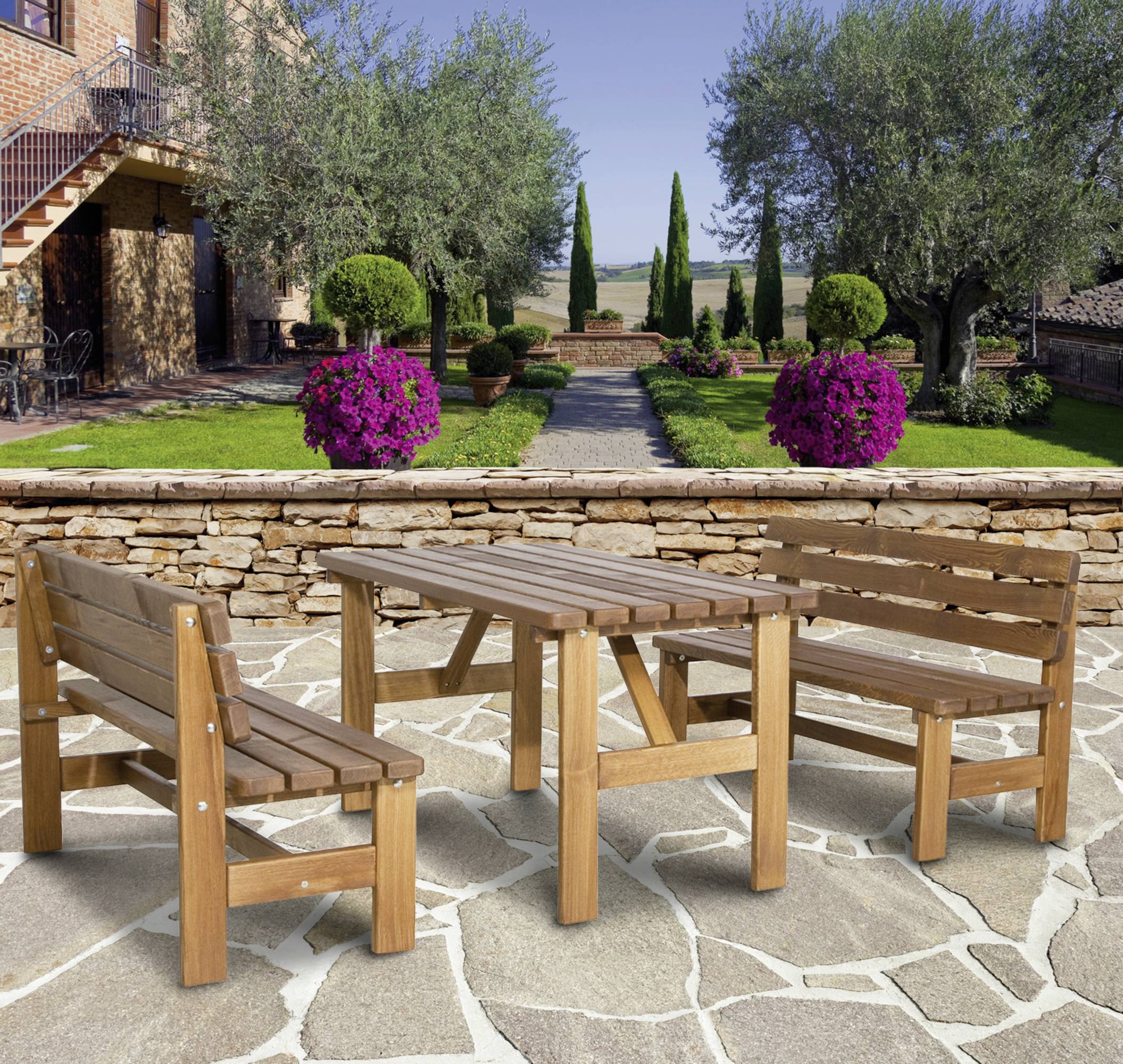 Drewniany komplet mebli na kamiennym tarasie przed starannie utrzymanym ogrodem z drzewami i fioletowymi kwiatami w doniczkach, w tle widoczny dom wiejski.