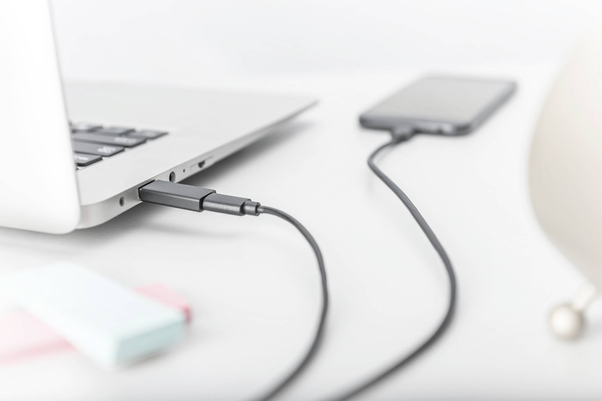 Smartfon jest podłączony do laptopa za pomocą kabla. Laptop leży na białym stole.