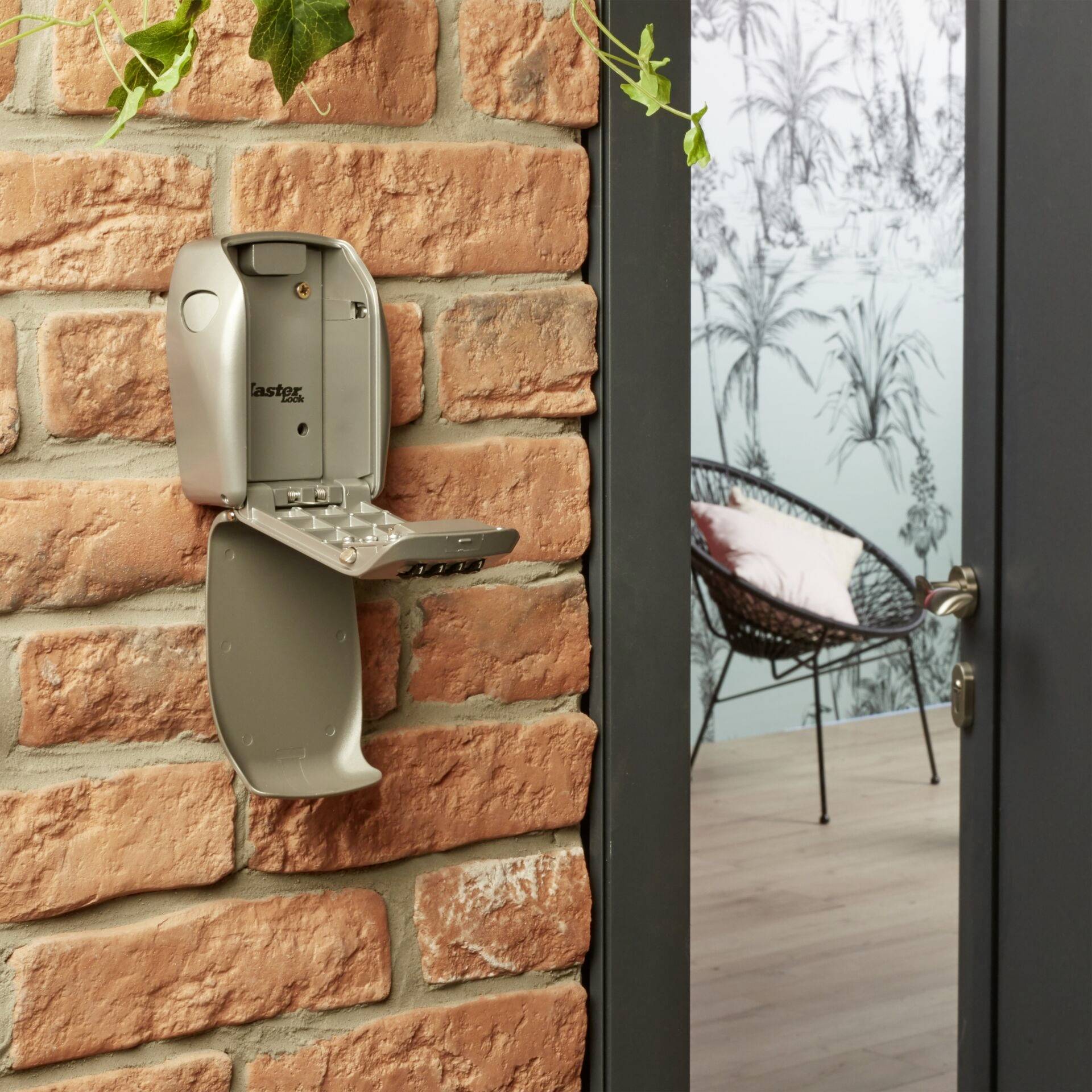 Skrzynka na klucze z otwartą pokrywą jest zamontowana na ścienie z cegły. Przez otwarte drzwi widać pokój z plecionym fotelem i tapetą na ścianie.