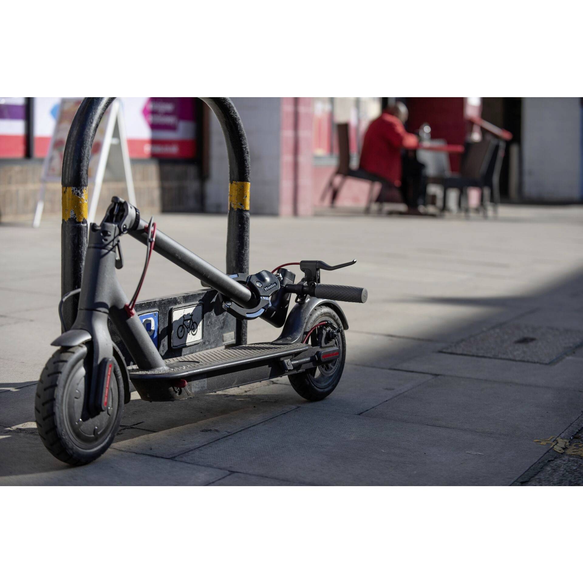 Złożony hulajnoga elektryczna stoi przy stojaku rowerowym na chodniku. W tle ktoś siedzi przy stole.