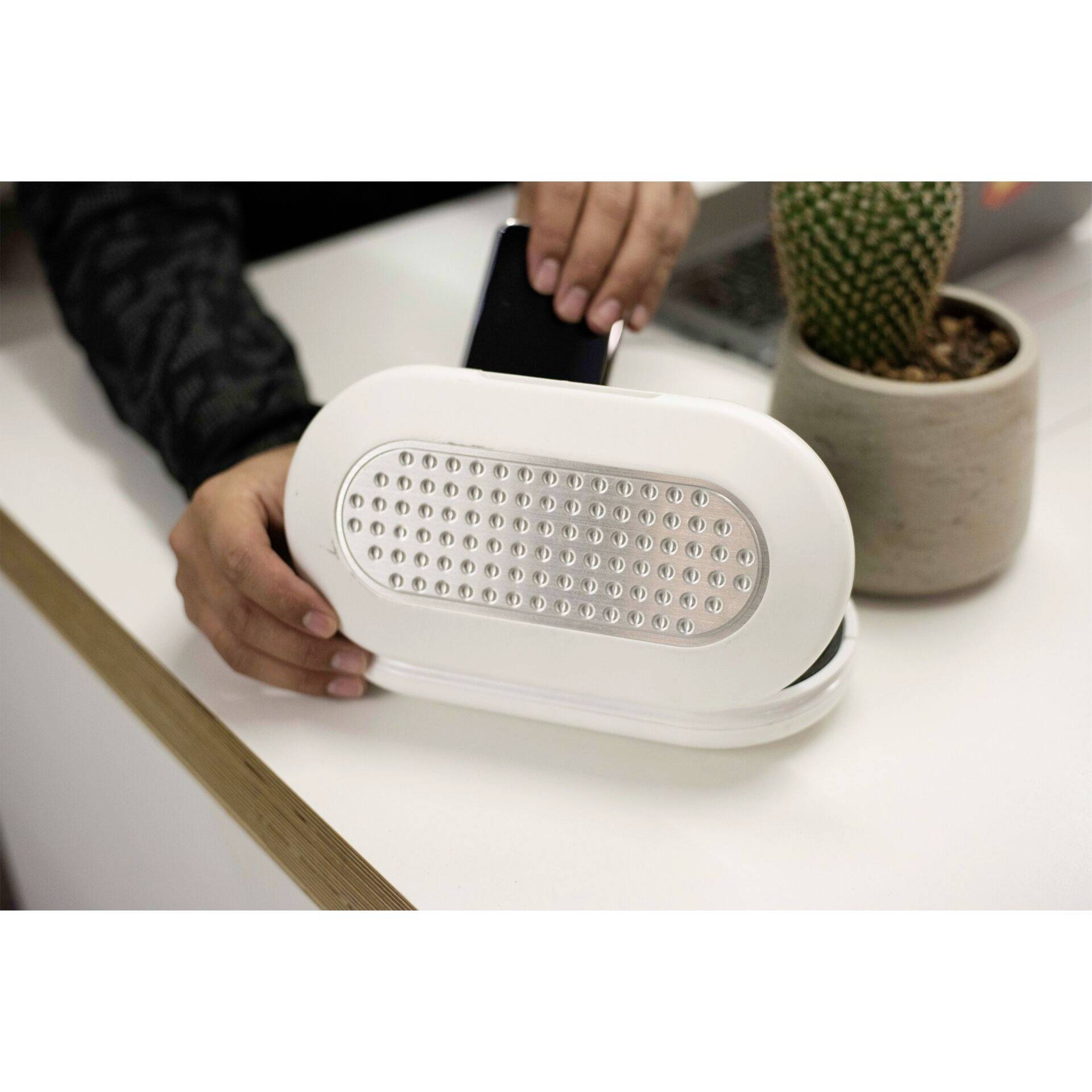 Osoba kładzie smartfon na białej ładowarce bezprzewodowej stojącej na stole obok małego kaktusa w doniczce.