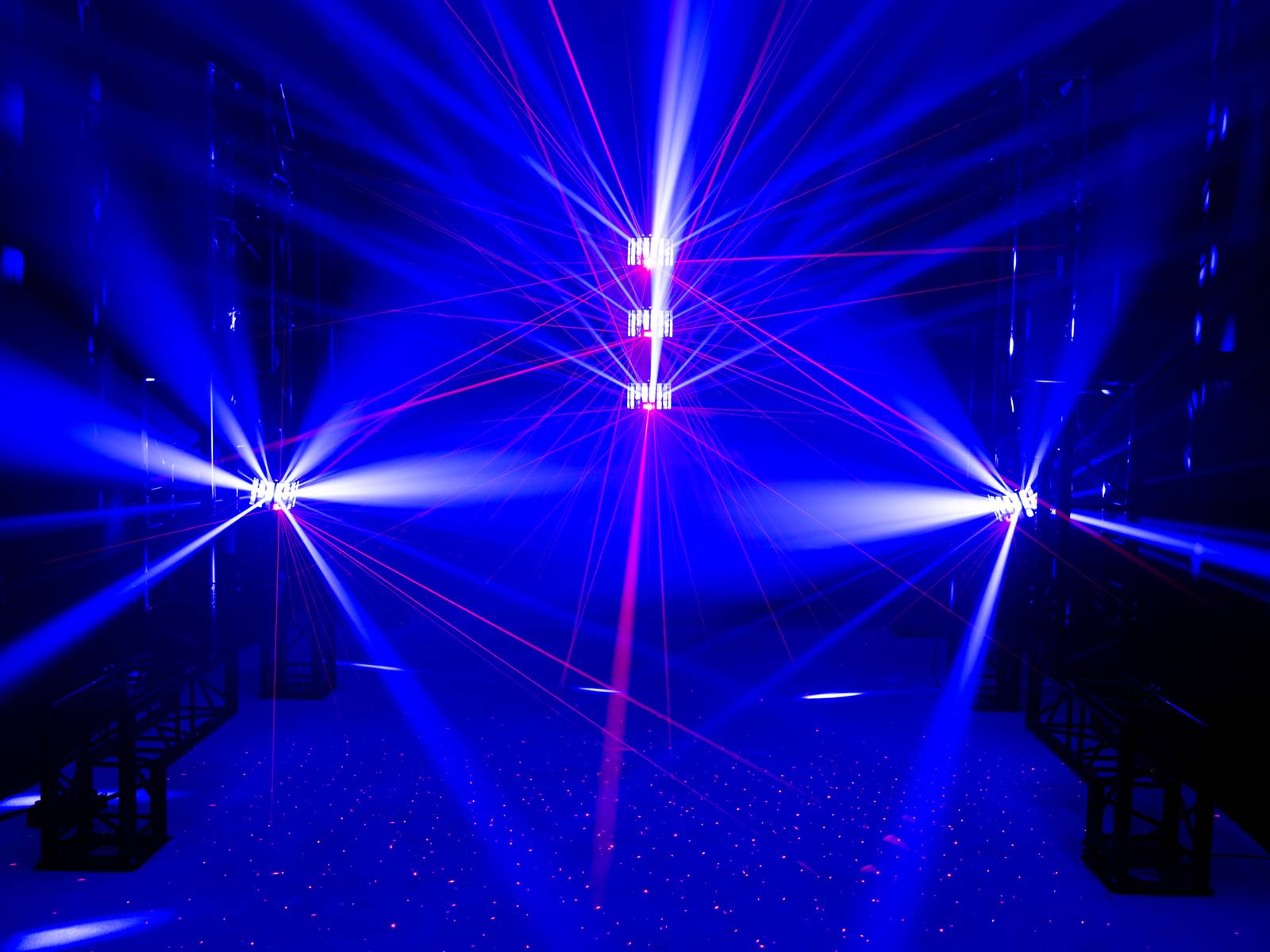 Niebieskie światło i pokaz laserowy w ciemnym pomieszczeniu, z skoncentrowanymi wiązkami światła wychodzącymi z centrum i z boku, tworzącymi dynamiczną scenografię.