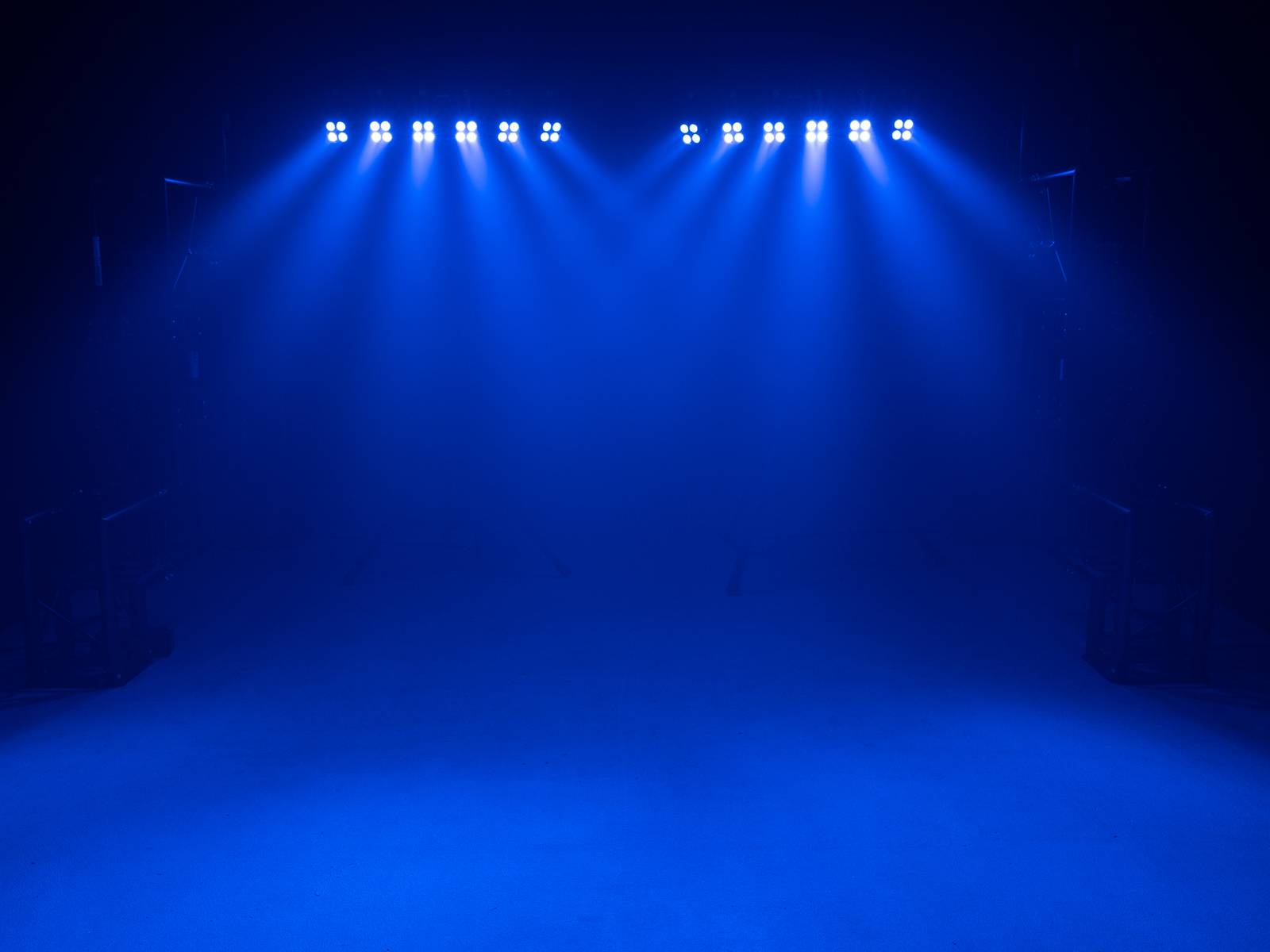 Kilka niebieskich świateł jasno rozbłyska, oświetlając ciemną scenę od góry. Atmosfera jest dramatyczna i pełna napięcia.