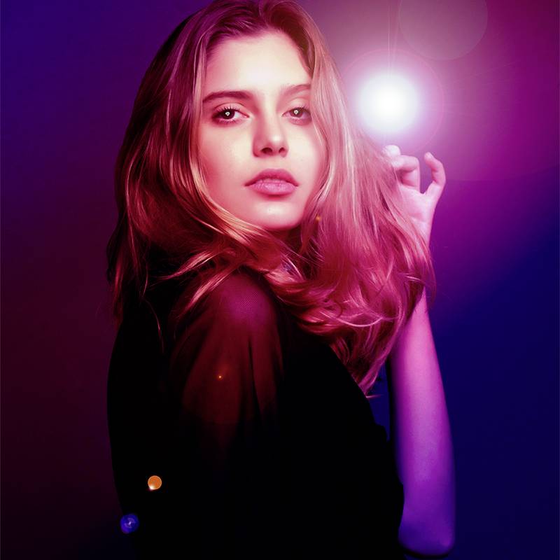 Osoba stojąca przed jasnym, kolorowym tłem, patrząca bezpośrednio w obiektyw. Ma długie włosy i jest ubrana na ciemno.