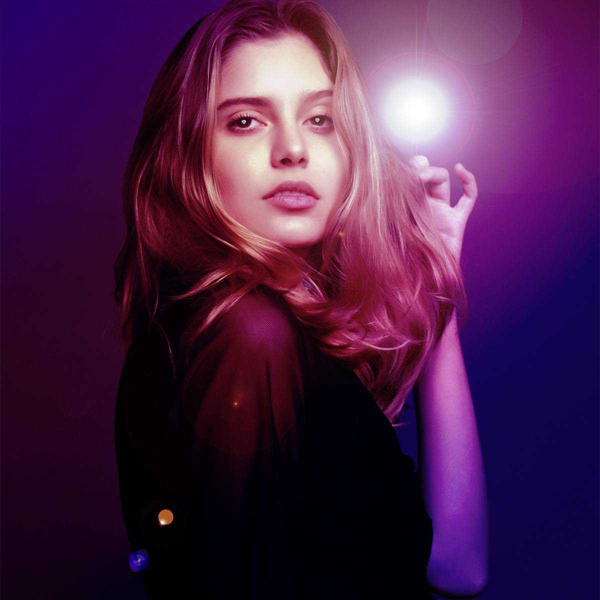 Osoba z długimi włosami pozuje przed fioletowym tłem, oświetlona jasnym światłem, które tworzy tajemniczą atmosferę.
