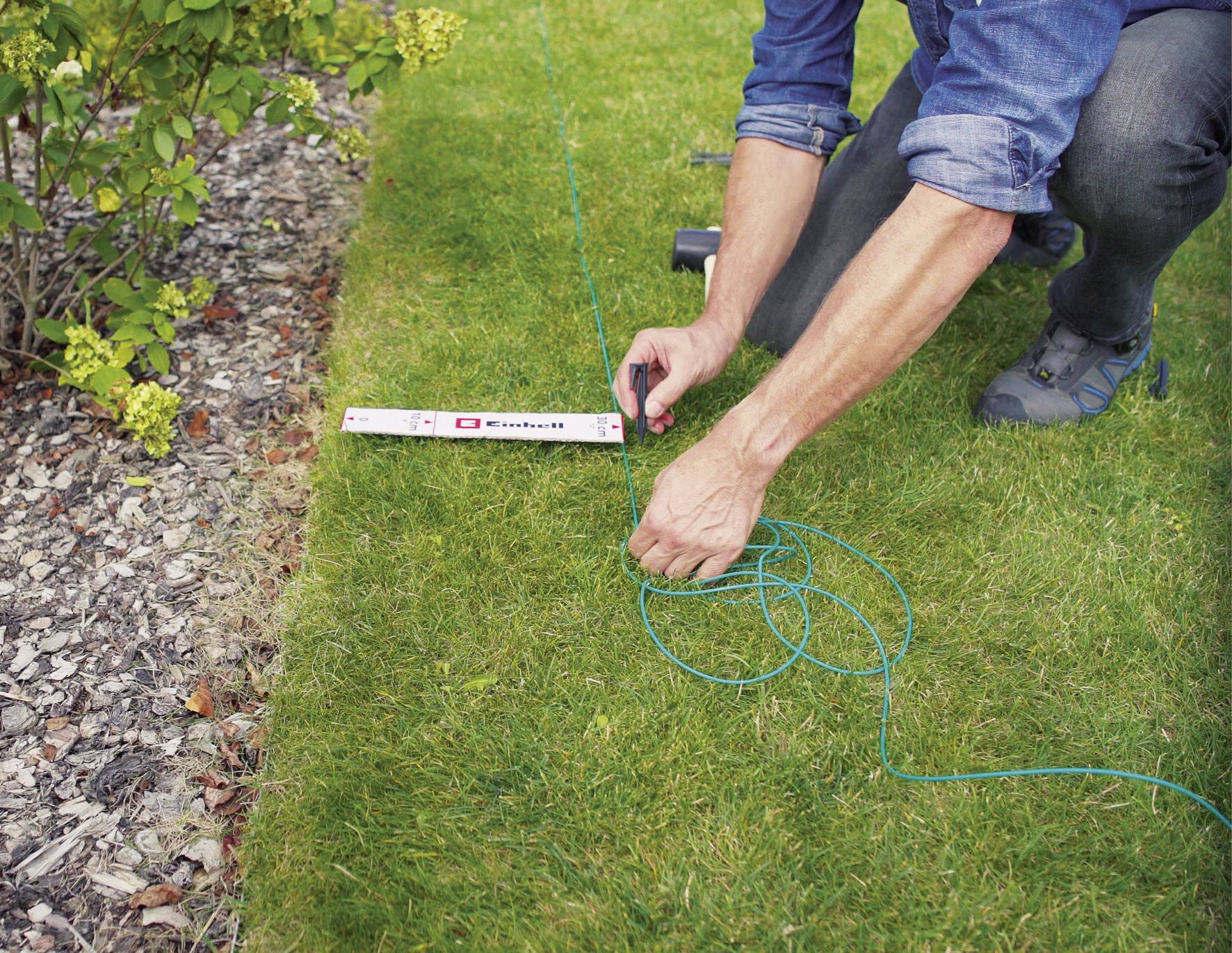 Osoba kładzie zieloną linkę wzdłuż trawnika, aby oznaczyć prostą linię. Widoczna jest miara taśmowa oraz krzewy.