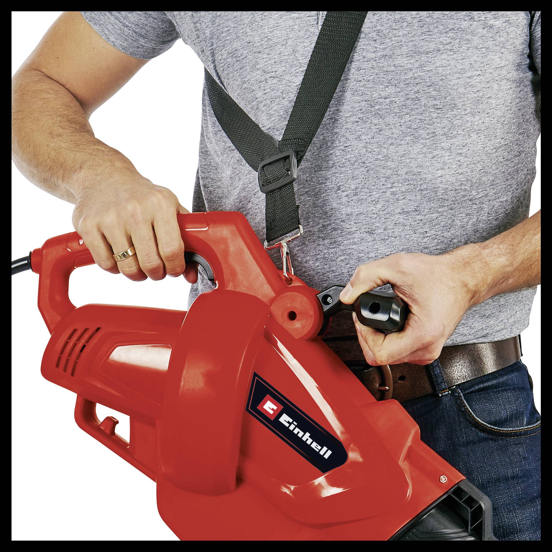 Osoba trzyma czerwone urządzenie dmuchające z logo Einhell, jest ubrana w szary t-shirt i niebieskie dżinsy. Urządzenie jest obsługiwane obiema rękami.