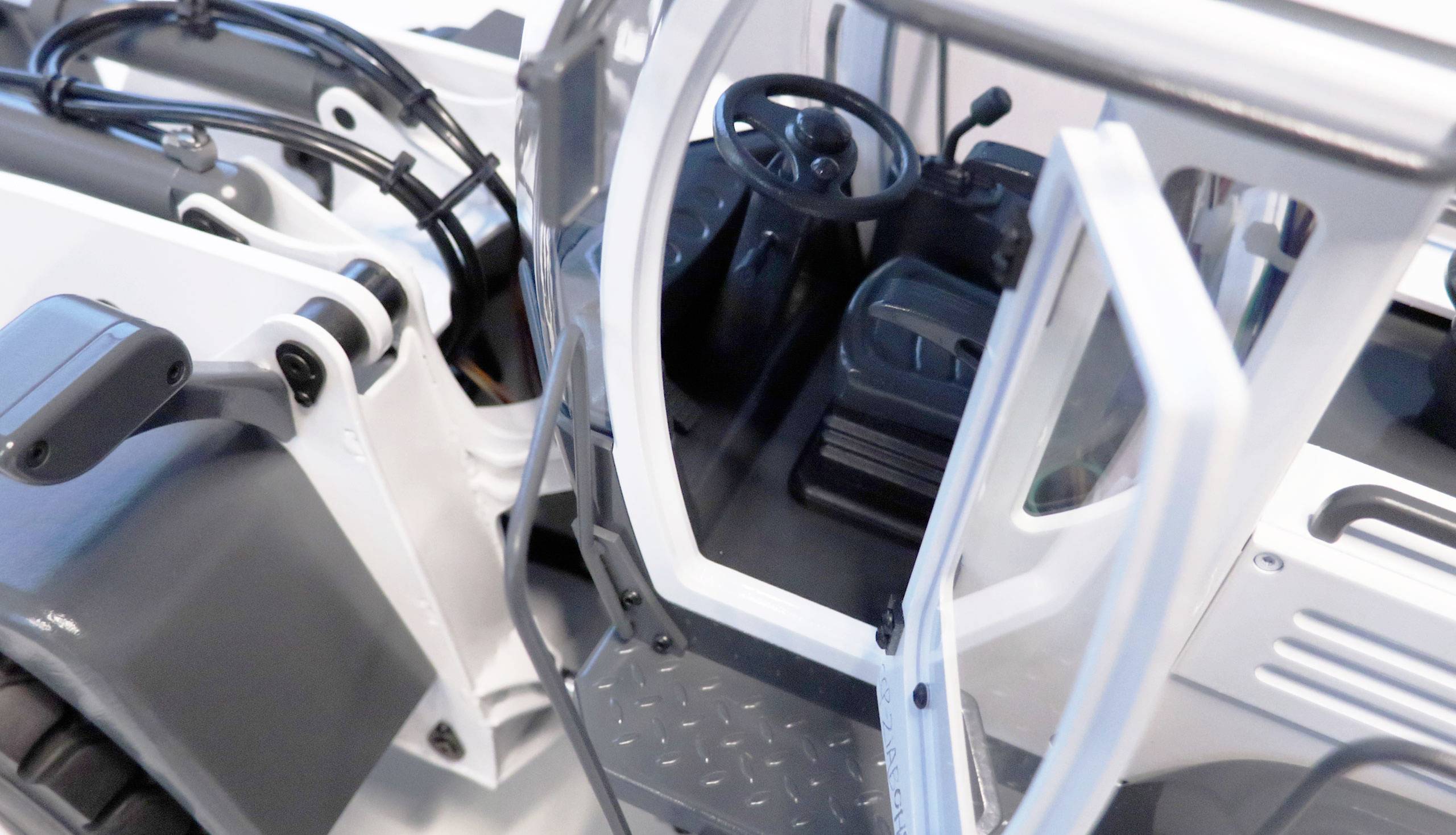 Widok wnętrza pojazdu przemysłowego z otwartą kabiną kierowcy, kierownicą, siedzeniem i dźwigniami sterowniczymi. Utrzymane głównie w białej kolorystyce.