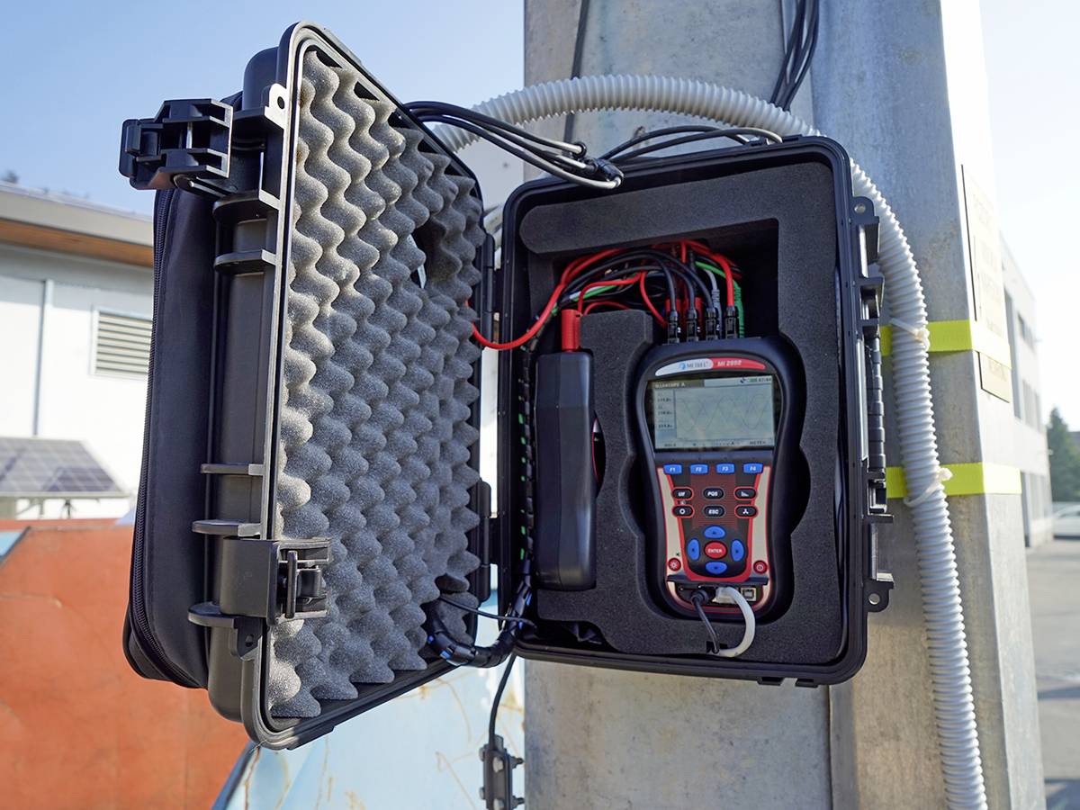 Otwarty kufer z urządzeniem pomiarowym podłączonym do kabli, zamontowany na słupie. W tle widoczne są budynki.