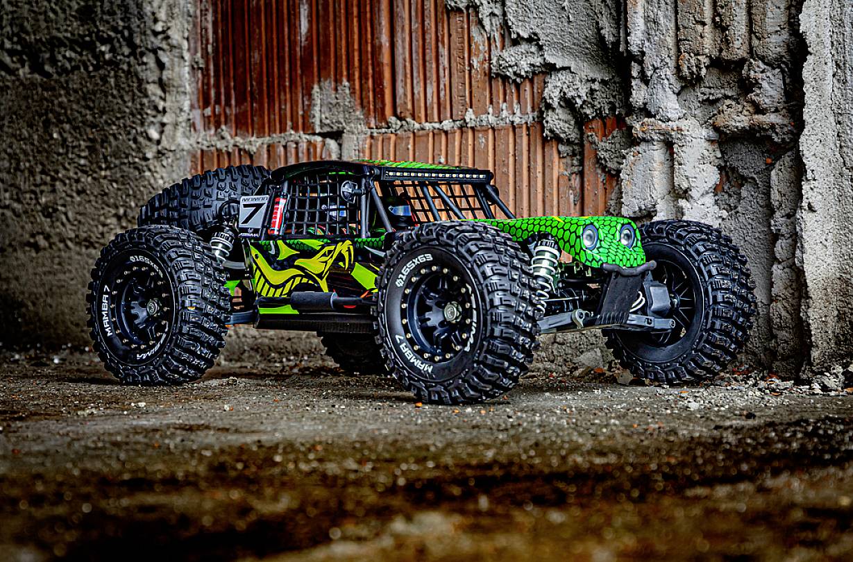 Zdalnie sterowany samochód z dużymi oponami i zieloną warstwą lakieru w kształcie węża stoi przed szorstką betonową ścianą w środowisku miejskim.