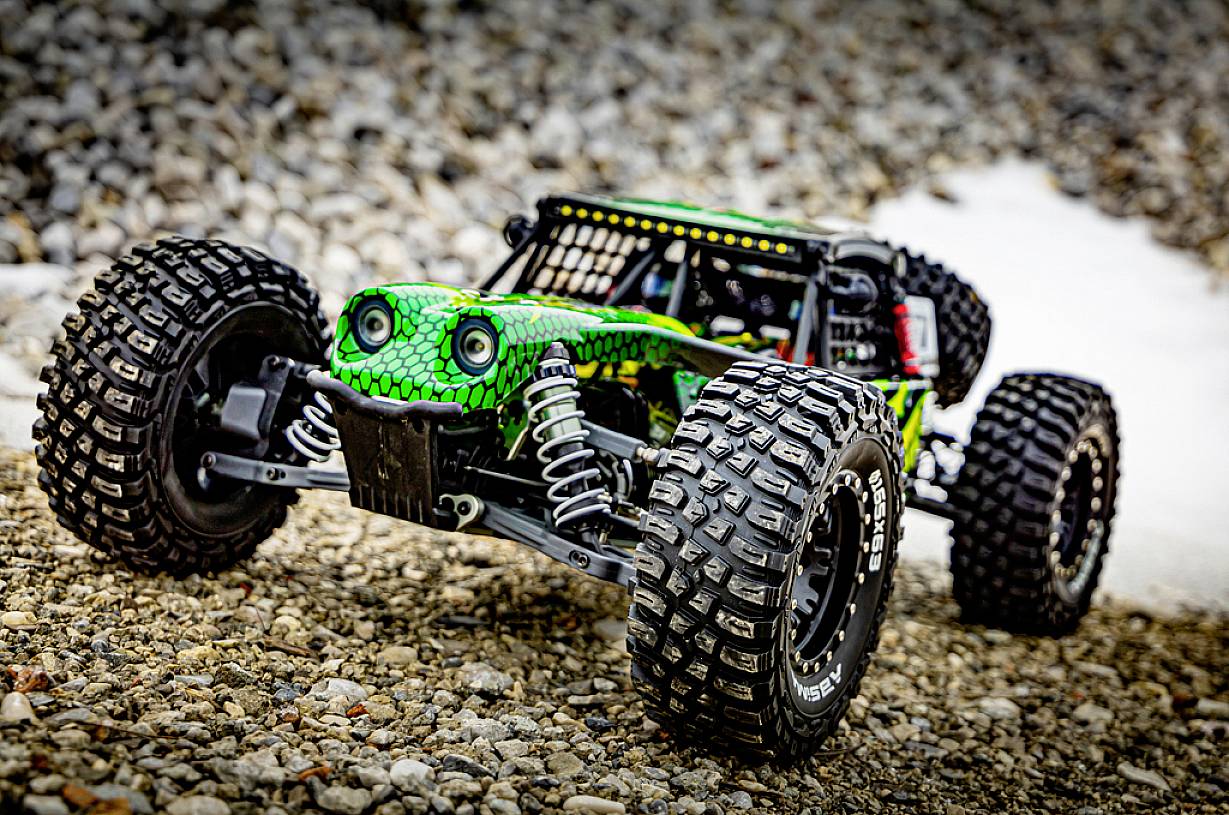 Zdalnie sterowane auto z zielonym wzorem w kształcie węża jedzie po żwirowej drodze. Widoczne są duże opony i zawieszenie.