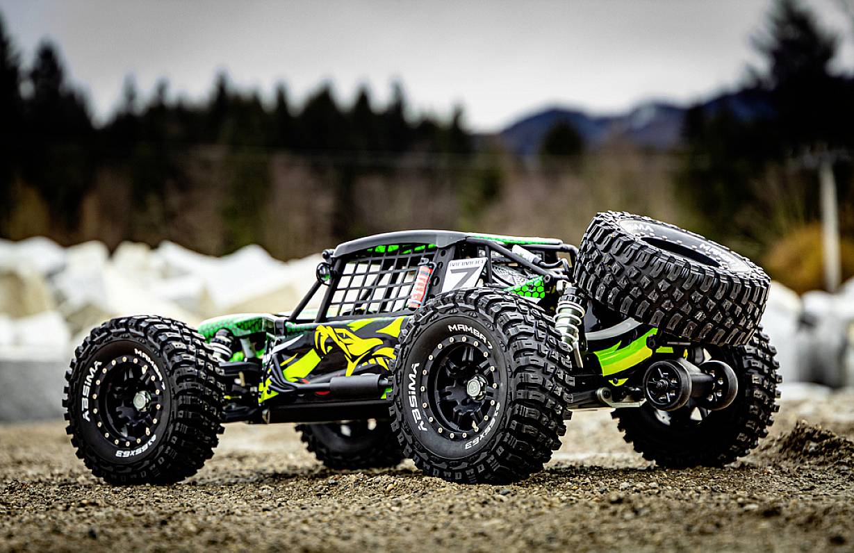 Zdalnie sterowany samochód terenowy z dużymi oponami i zielonymi akcentami stoi na nieutwardzonej drodze przed zalesionymi wzgórzami.