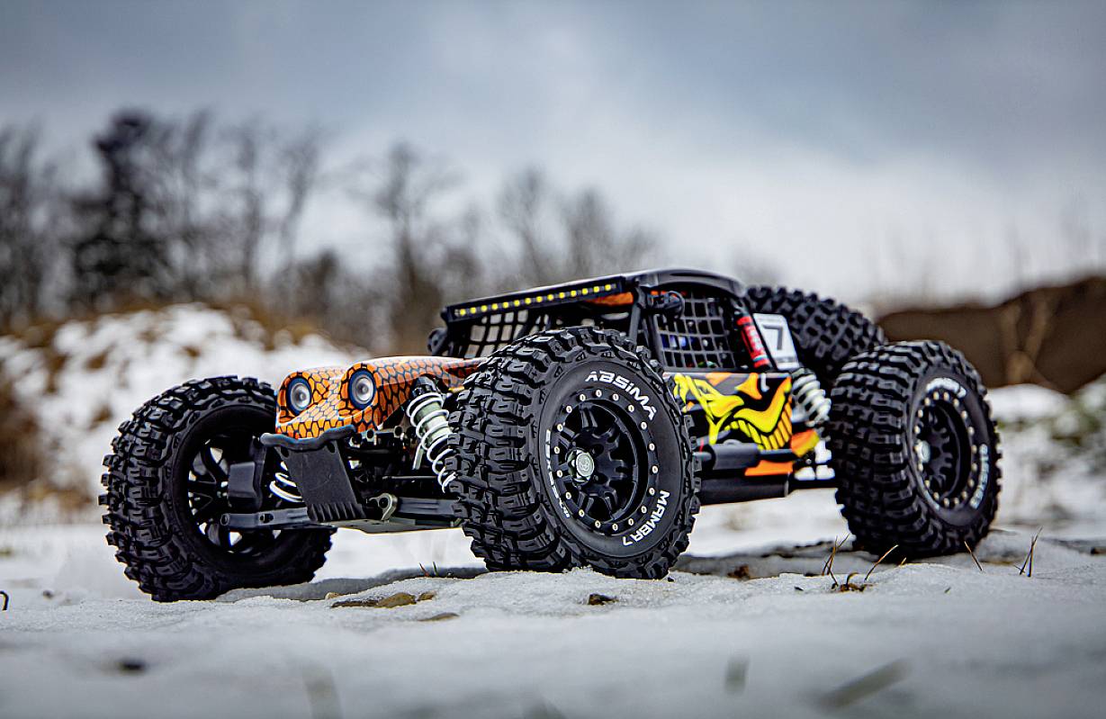 Zdalnie sterowany samochód z dużymi oponami jedzie po zaśnieżonej przestrzeni. W tle drzewa rysują się niewyraźnymi zarysami.