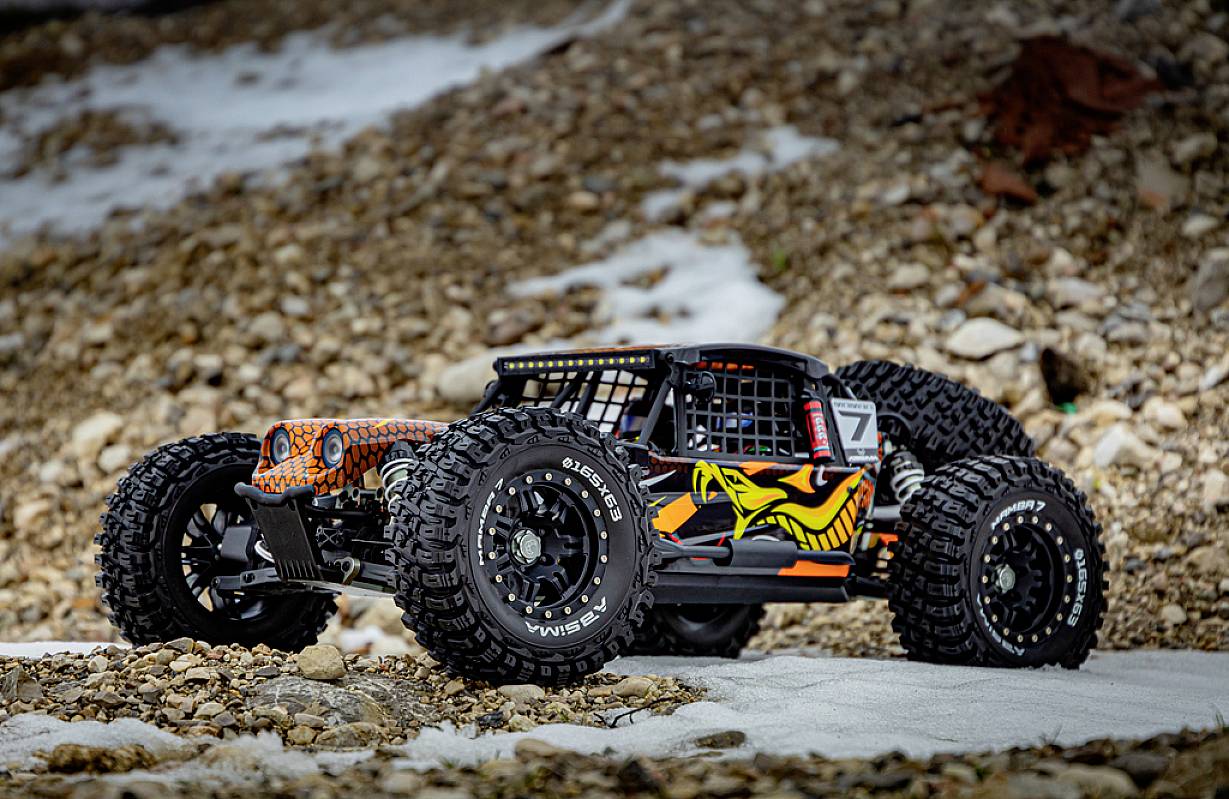 Zdalnie sterowany samochód z grubym bieżnikiem opon stoi na kamienistym podłożu. Jest czarno-pomarańczowy z żółtym wzorem węża.