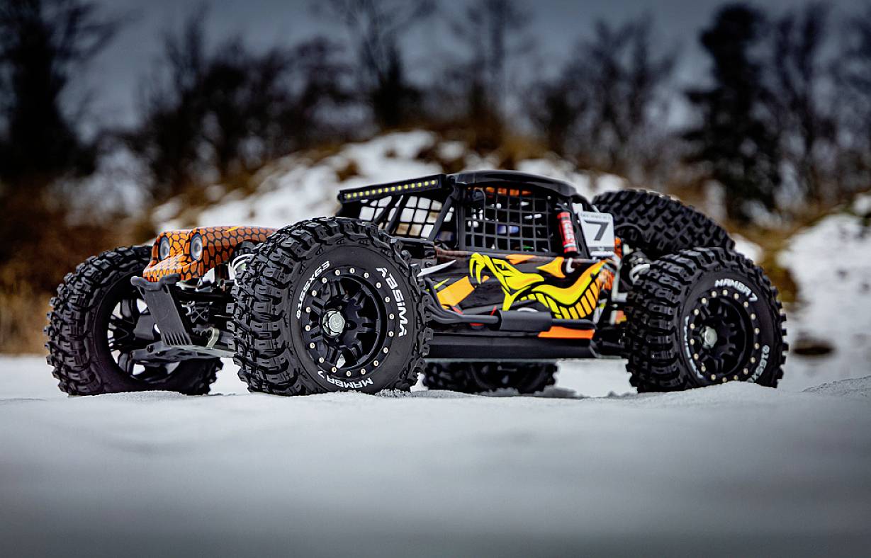 Zdalnie sterowany samochód na śniegu, czarno-pomarańczowy z rzucającym się w oczy projektem graficznym na bokach. Grube opony i ośnieżone tło.