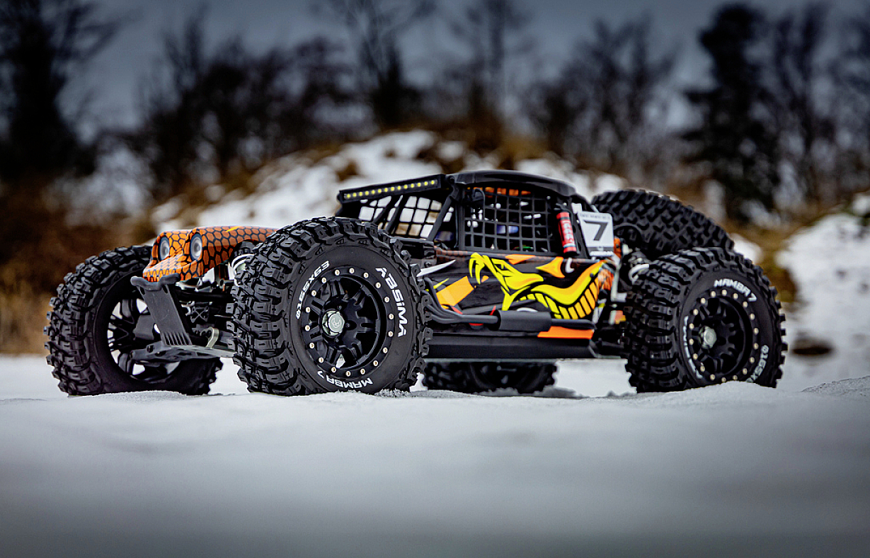 Zdalnie sterowany samochód na śniegu, czarno-pomarańczowy z rzucającym się w oczy projektem graficznym na bokach. Grube opony i ośnieżone tło.