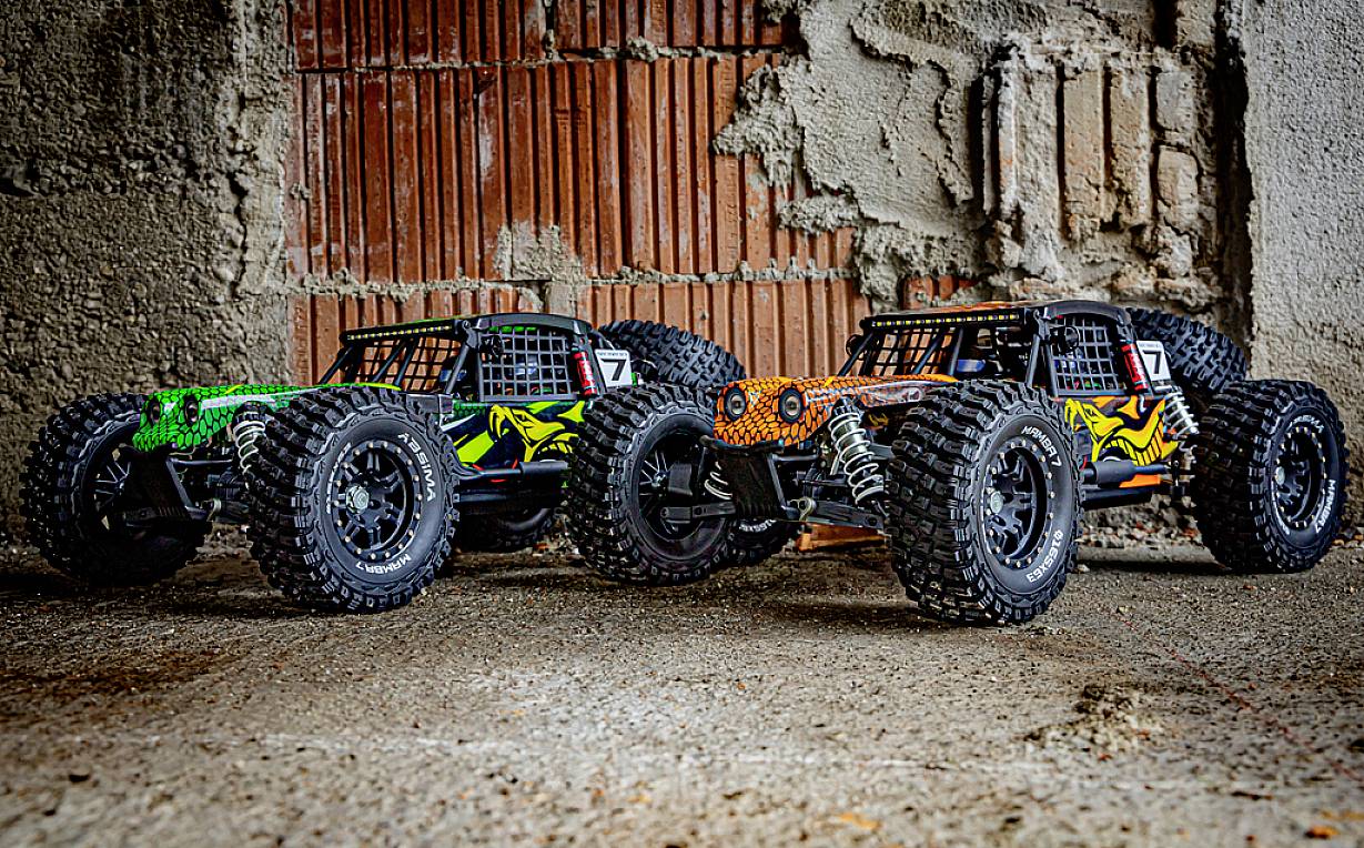 Trzy kolorowe zdalnie sterowane samochody terenowe są umieszczone obok siebie na betonowej podłodze przed surową ścianą z cegły.