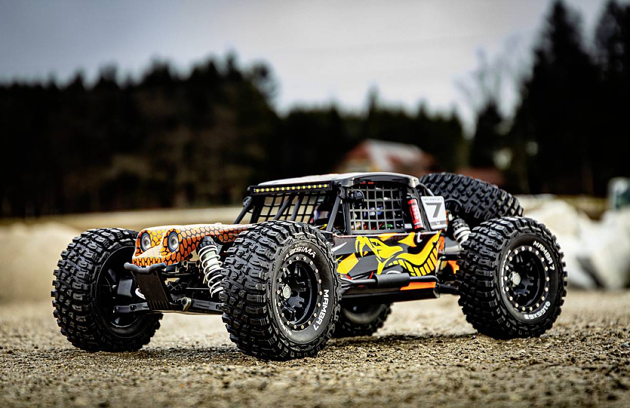 Zdalnie sterowany samochód terenowy z rzucającymi się w oczy grafikami i dużymi oponami do jazdy w terenie stoi na żwirowym podłożu na tle lasu.