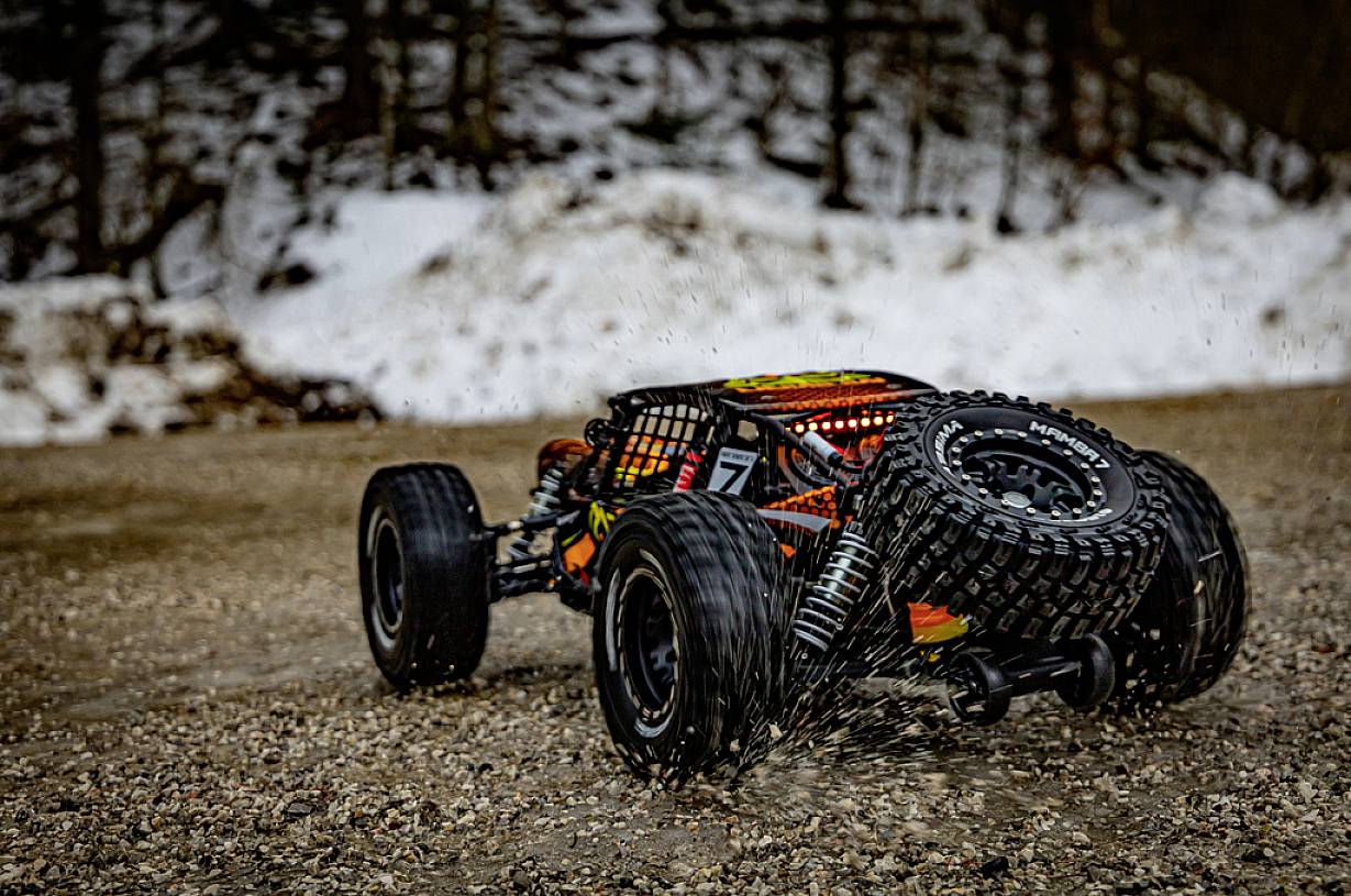 Zdalnie sterowany samochód jedzie po zaśnieżonej żwirowej drodze. W tle widoczne są drzewa i śnieg.