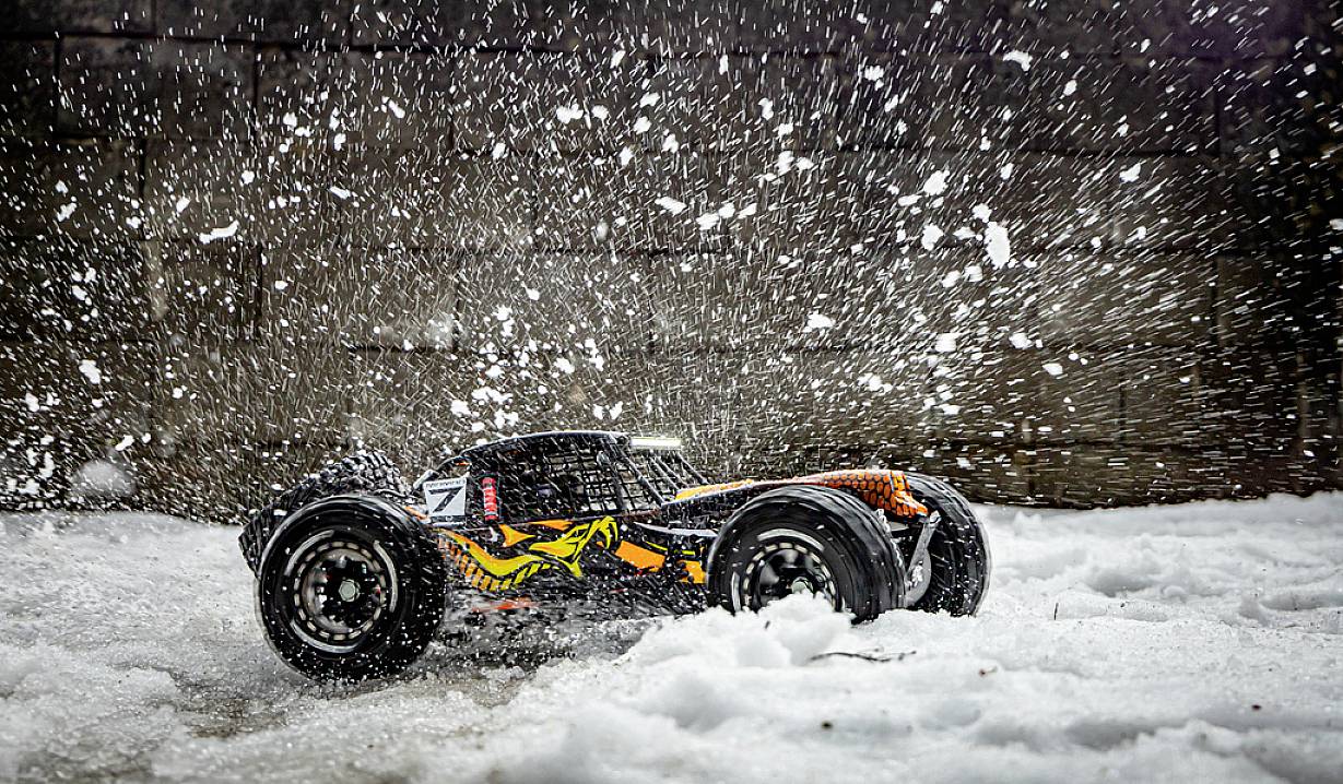 Zdalnie sterowane auto jedzie przez śnieg, podnosząc płatki śniegu w powietrze. W tle widnieje szara ściana.