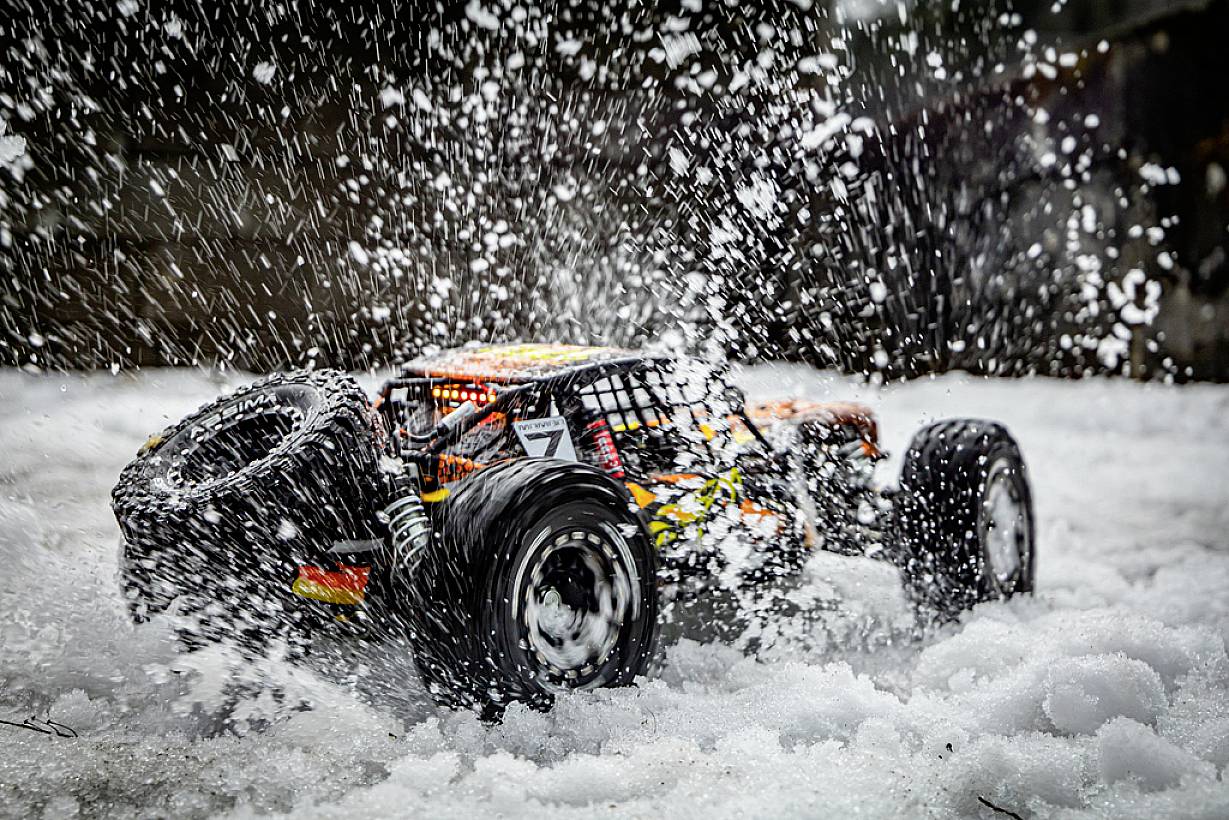 Zdalnie sterowane auto rozpryskuje śnieg. Porusza się dynamicznie, otoczone płatkami śniegu, co podkreśla jego prędkość i zwrotność.