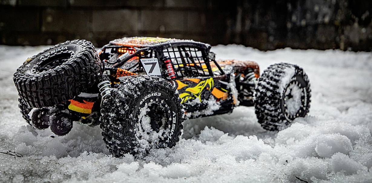 Zdalnie sterowany samochód z dużymi kołami jedzie przez śnieg, a jego karoseria jest w kolorach pomarańczowym, żółtym i czarnym.