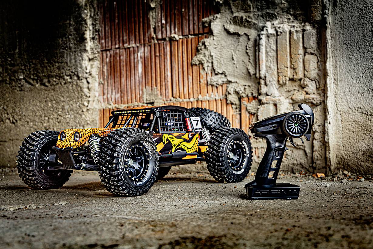 Zdalnie sterowane auto z dużymi oponami stoi na betonowej podłodze obok pilota, przed rustykalną ścianą z cegły.