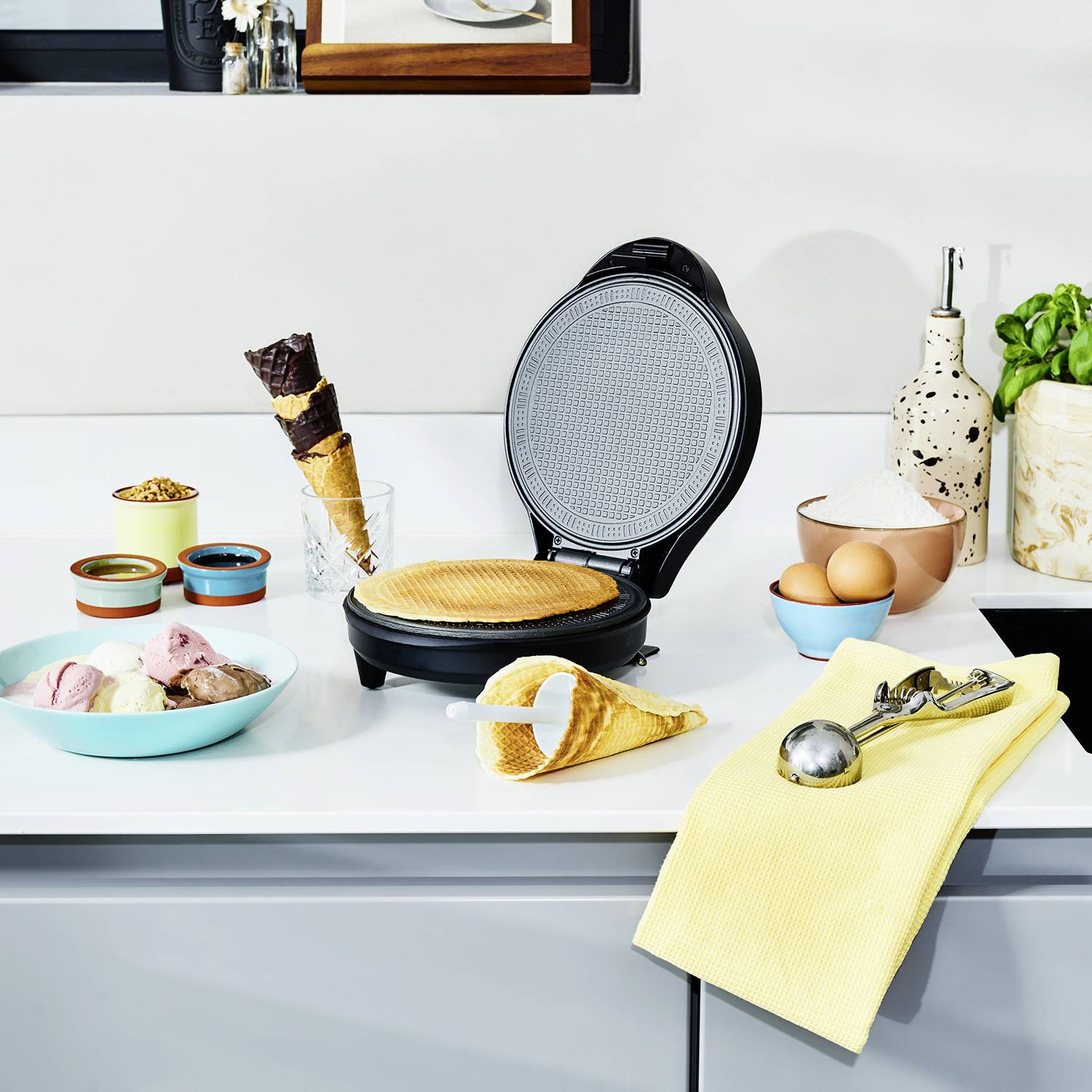 Żelazko do gofrów na blacie kuchennym, otoczone rożkami, lodami, goframi oraz składnikami takimi jak jajka i mąka.