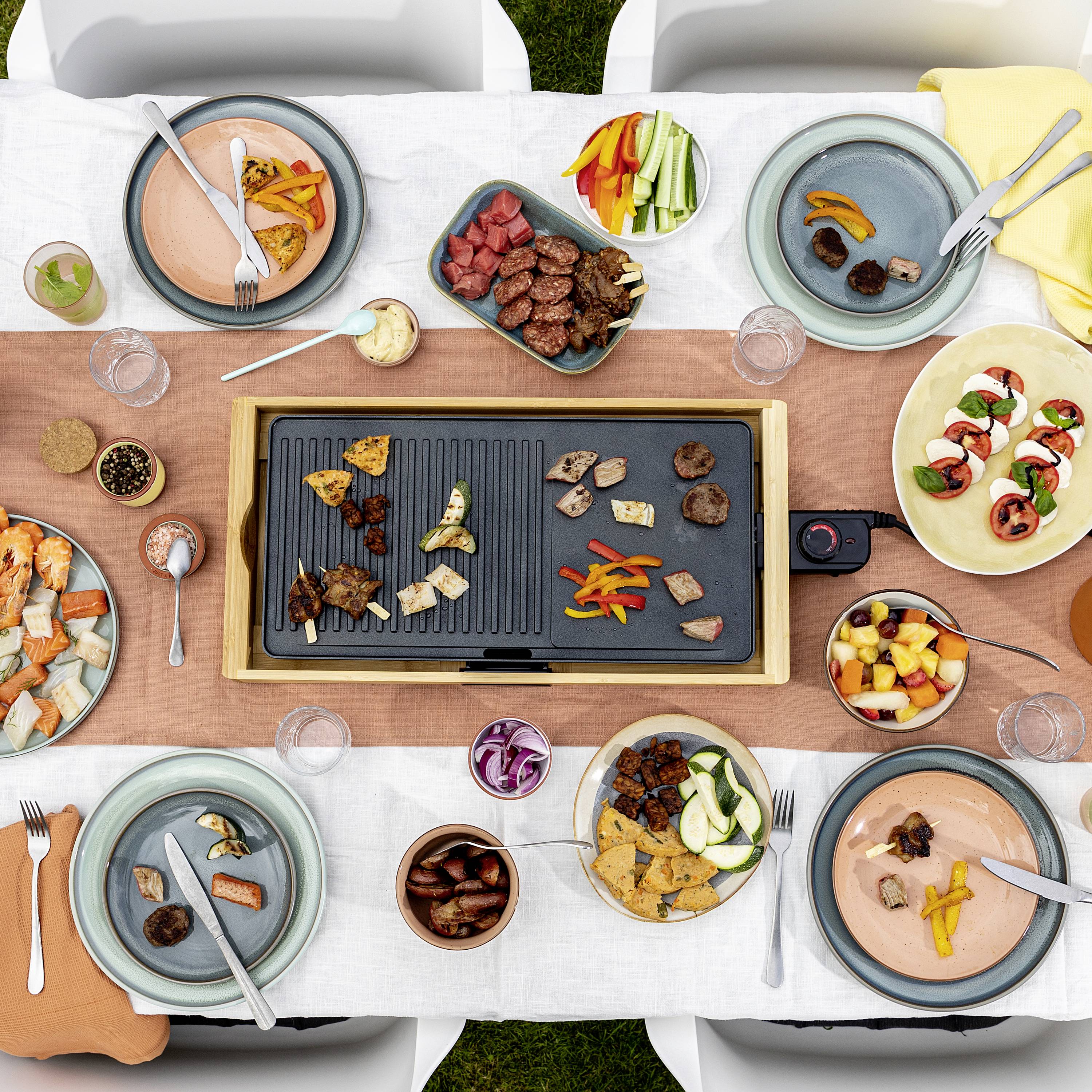 Scena przy stole z elektryczną płytą grillową pośrodku, otoczoną różnymi talerzami z mięsem, warzywami i owocami.