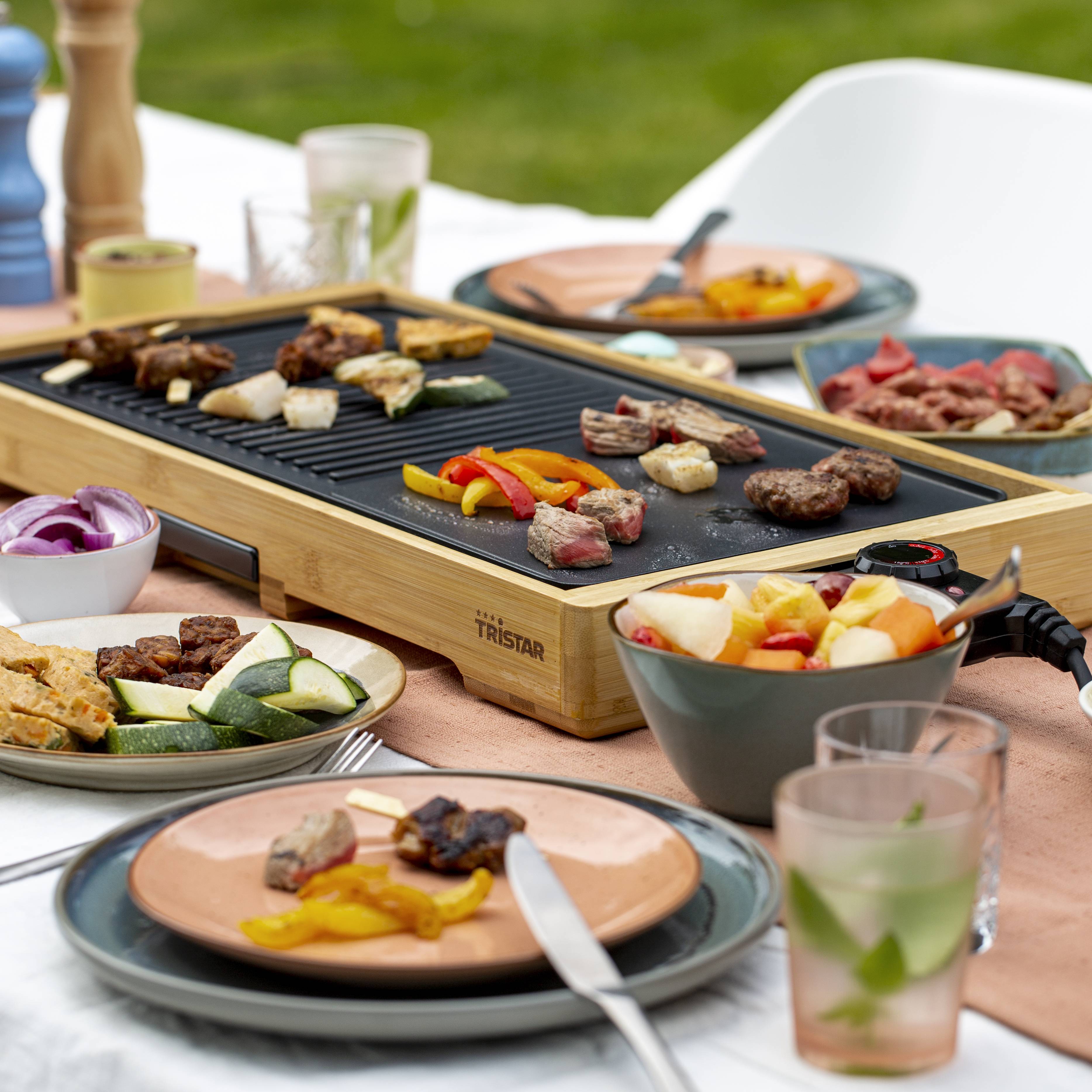 Stolik na świeżym powietrzu z płytą grillową, na której piecze się mięso i warzywa. Talerze z jedzeniem i napoje są przygotowane.