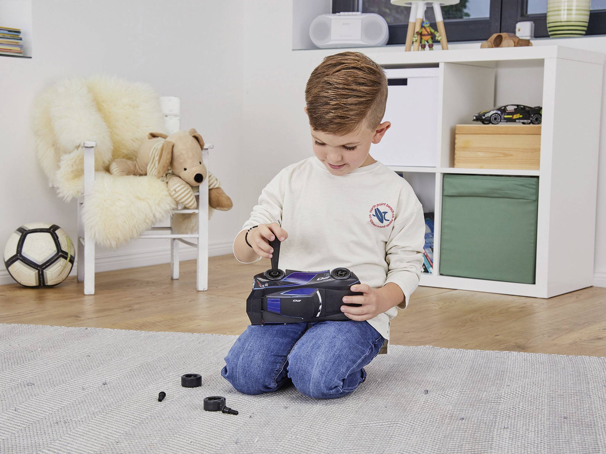 Chłopiec siedzi na podłodze i bawi się samochodem zdalnie sterowanym. Wokół niego rozrzucone są części samochodowe, a w tle widać zabawki.