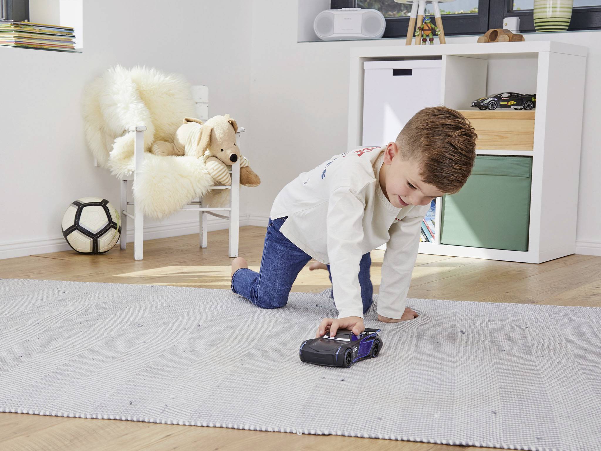 Chłopiec wesoło bawi się zabawkowym samochodem na dywanie w jasnym pokoju dziecięcym. W tle widać regał z zabawkami.