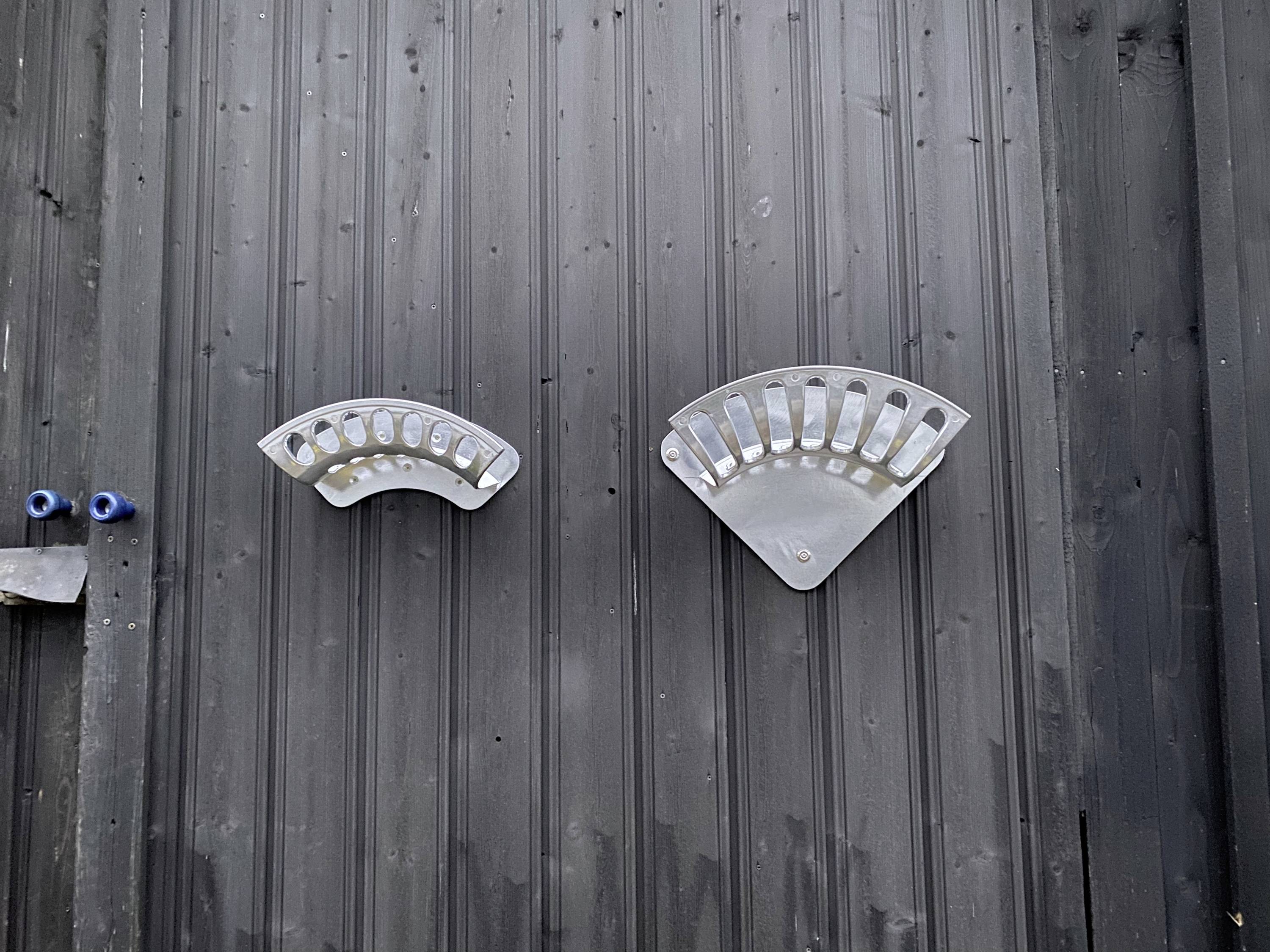 Dwie srebrne pokrywy wentylacyjne na ciemnej ścianie, rozmieszczone symetrycznie obok siebie.