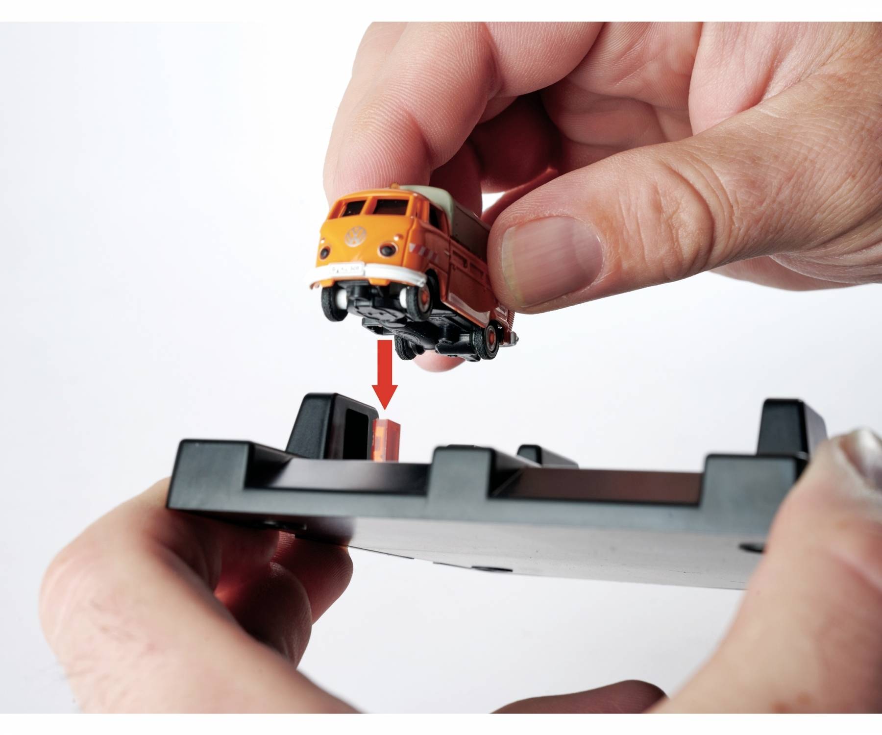 Dwie ręce umieszczają małe autko modelarskie na czarnej platformie. Czerwona strzałka wskazuje na gniazdo na platformie.