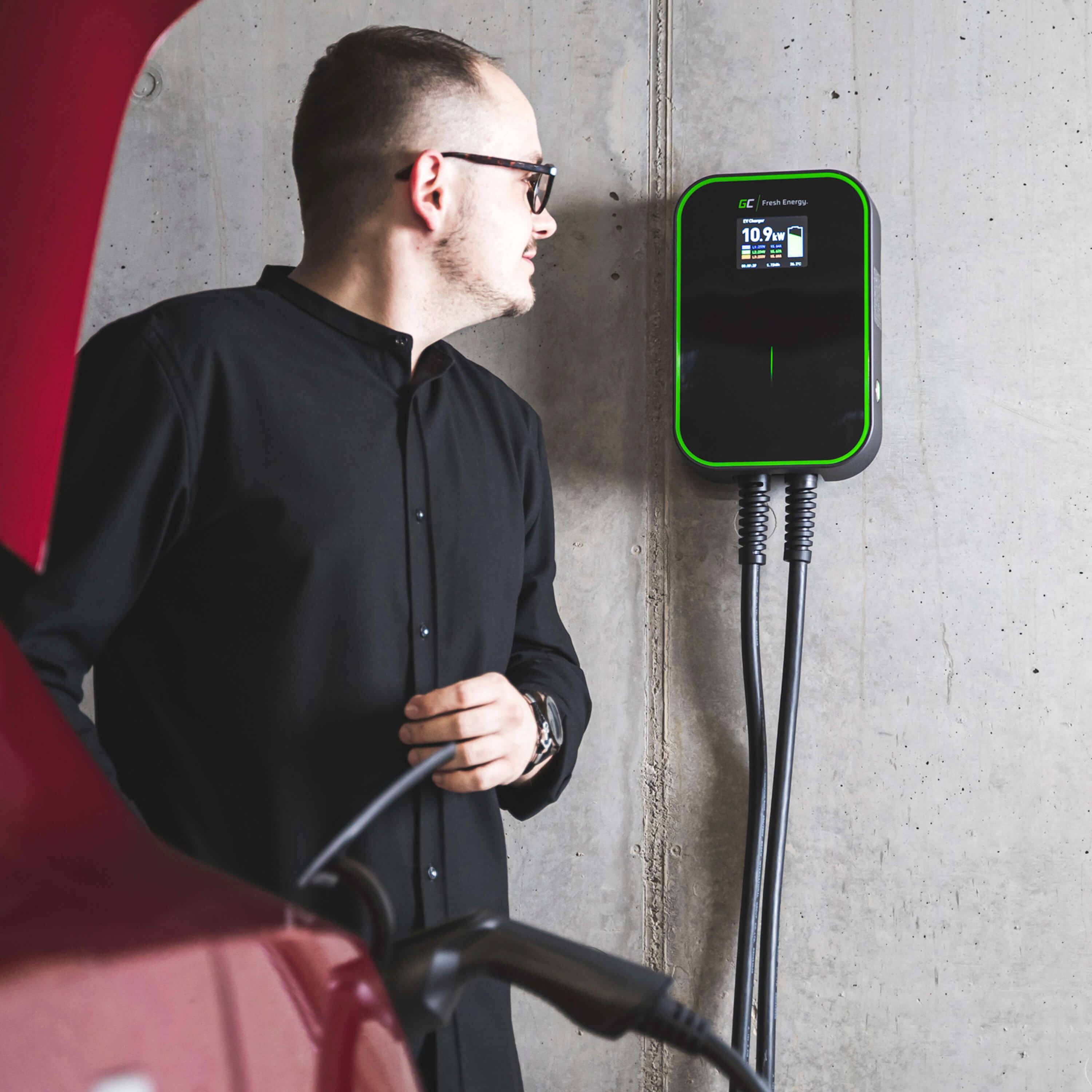 Mężczyzna w okularach przeciwsłonecznych stoi przed stacją ładowania samochodów elektrycznych zamocowaną na ścianie i obsługuje ją.