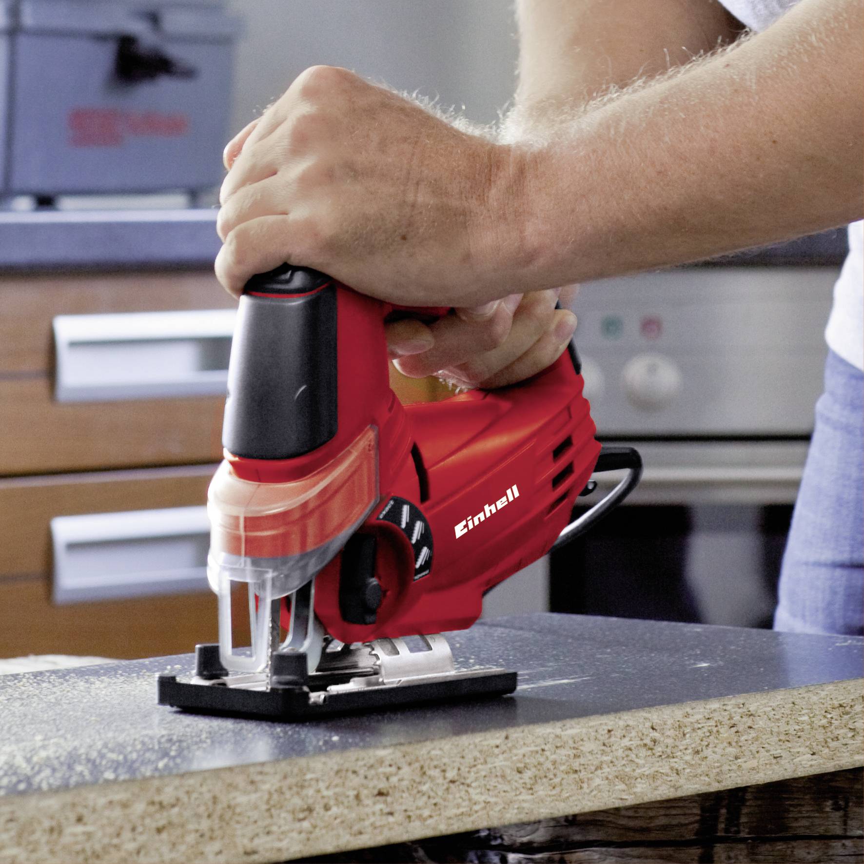 Osoba używa czerwonej elektrycznej wyrzynarki do cięcia kawałka drewna w warsztacie lub kuchni.