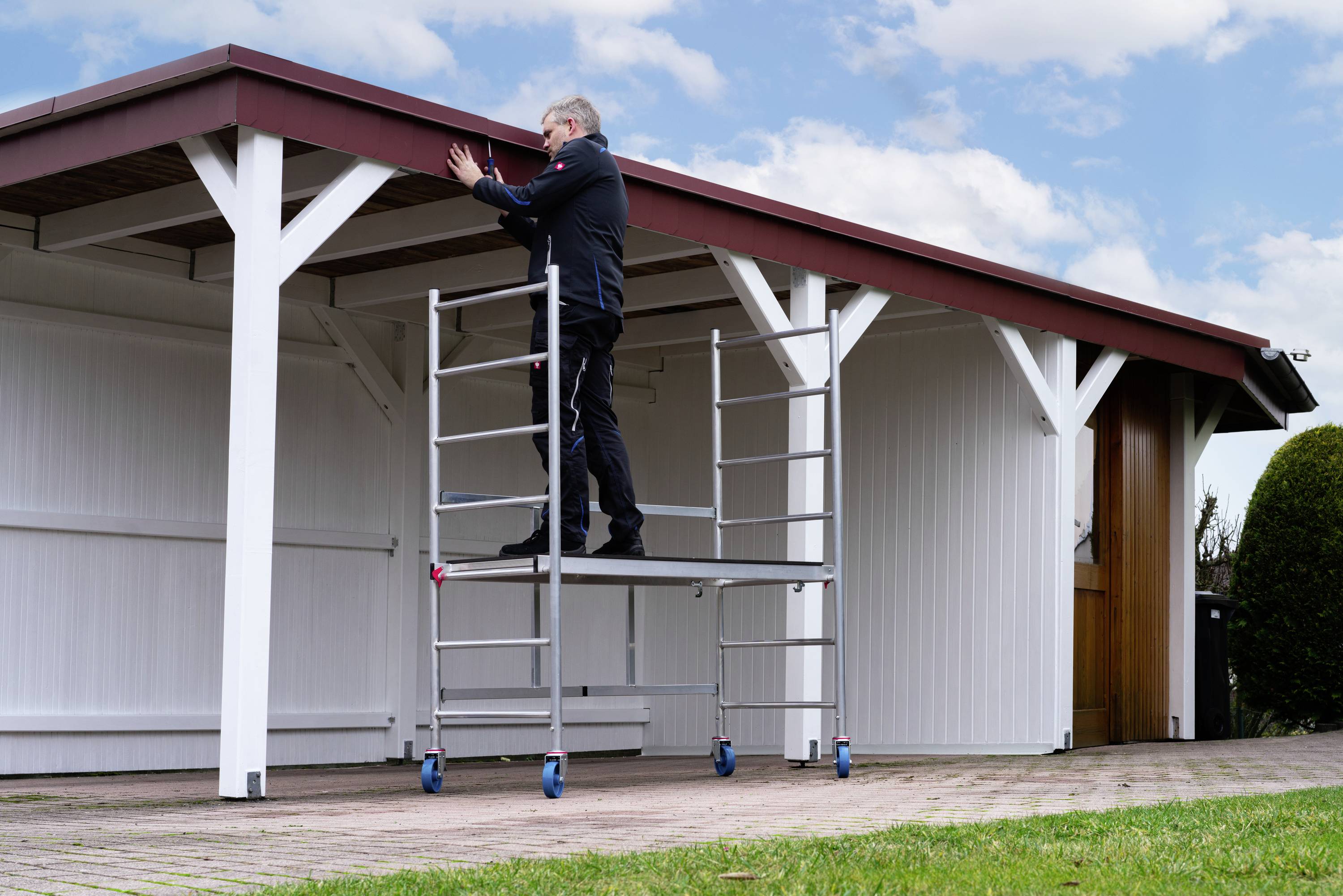 Mężczyzna stoi na rusztowaniu jezdnym i pracuje przy dachu białej drewnianej konstrucji obok garażu, na wolnym powietrzu.