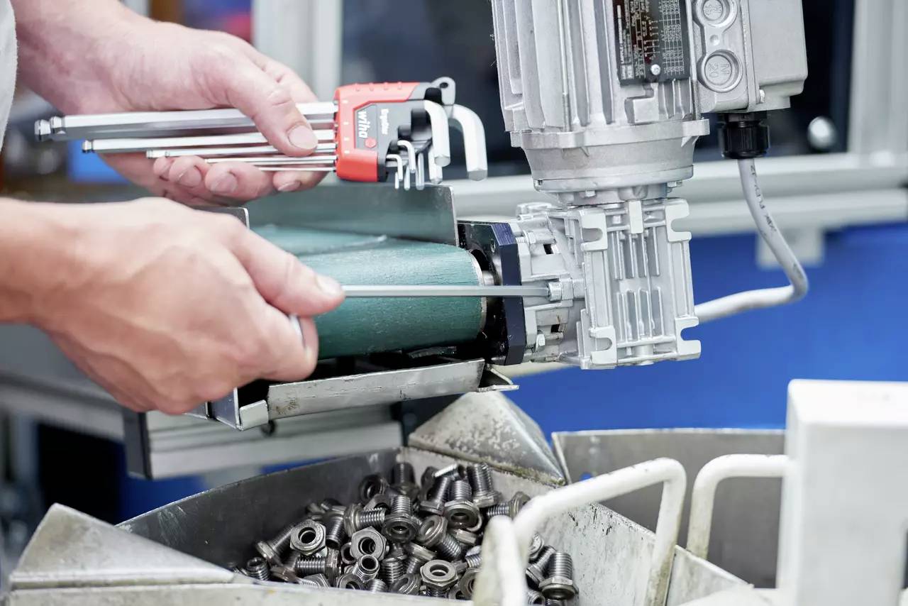 Osoba używa narzędzia do pomiaru elementów w środowisku maszynowym, z metalowymi częściami zgromadzonymi poniżej, co wskazuje na proces produkcyjny.