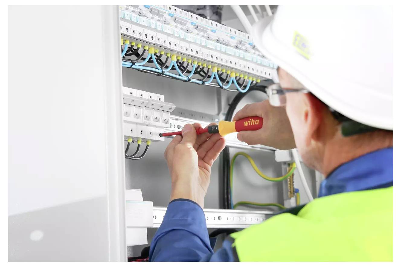 Elektryk w kasku i kamizelce odblaskowej używa śrubokrętu do regulacji okablowania w panelu elektrycznym.