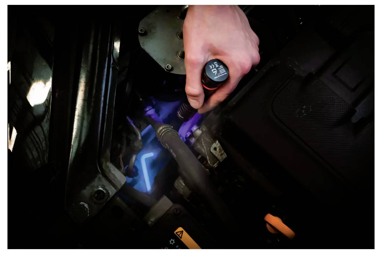 Ręka trzymająca sondę olejową nad silnikiem samochodu, sprawdzająca poziom oleju. Widoczne części silnika, oświetlone neonowym niebieskim światłem.