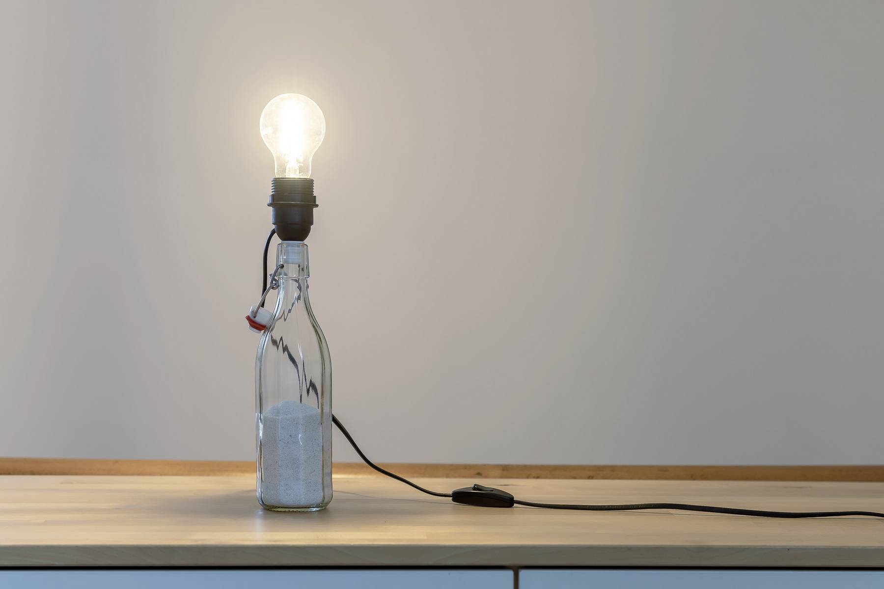 Świecąca żarówka spoczywa na szklanej butelce wypełnionej piaskiem. Przewód z włącznikiem łączy lampę z zasilaniem elektrycznym.