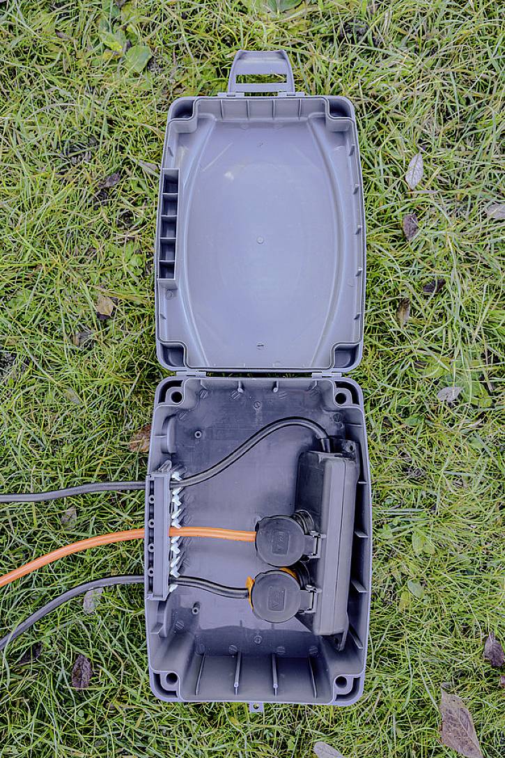 Otwarta szara puszka przyłączeniowa z widocznymi złączami kabli i dwiema gniazdkami elektrycznymi, umieszczona na pokrytej trawą ziemi.