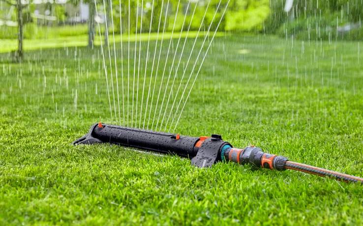 Osoba podłącza pomarańczowo-szary złącznik węża do metalowego kranu na dworze, na rozmytym zielonym tle ogrodu.